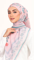 A woman wearing a light pink hijab with a blue floral pattern and a thin teal border, paired with a white top against a plain white background.