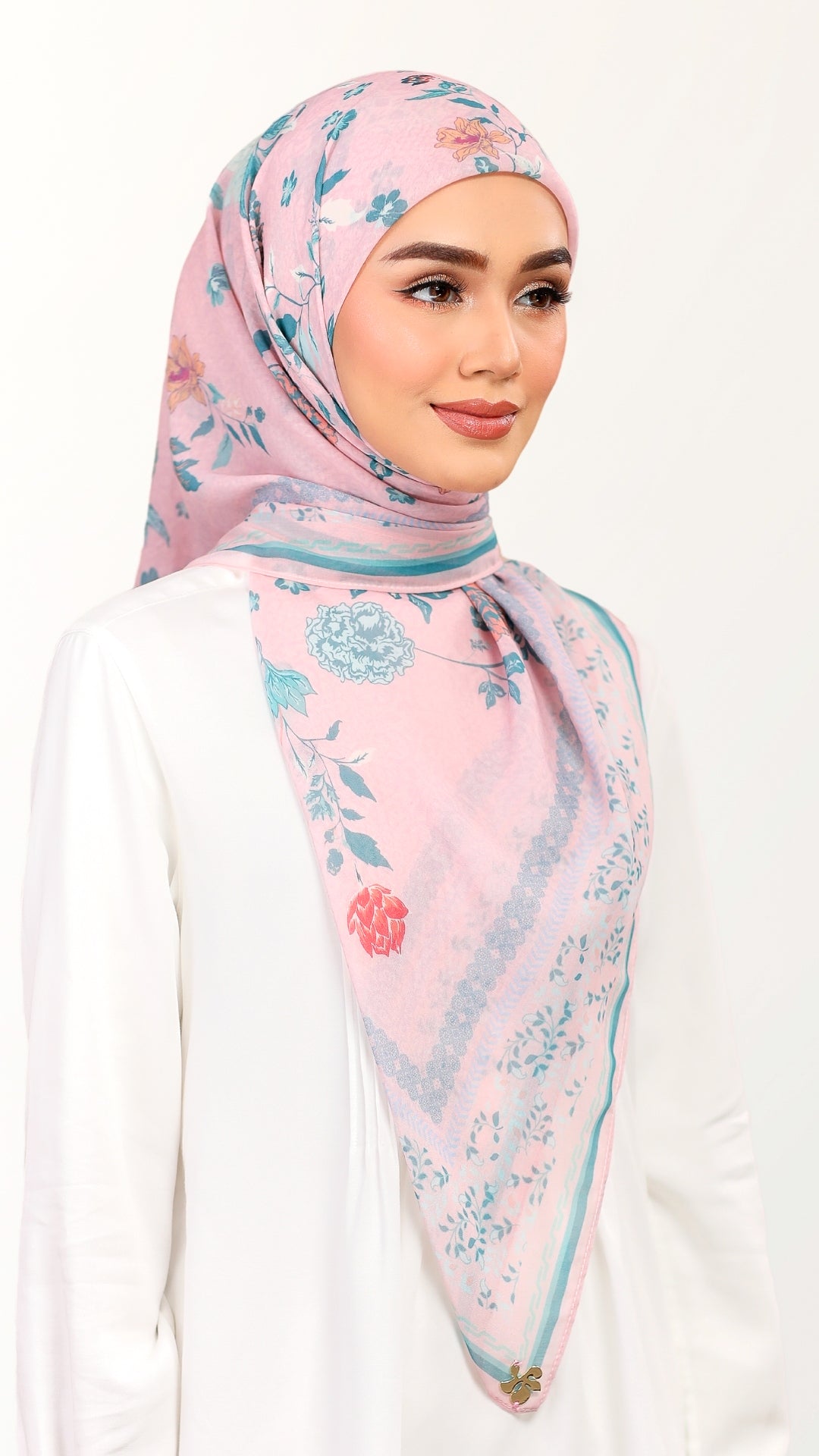 A woman wearing a light pink hijab with a blue and red floral print, styled with a white long-sleeve top against a plain white background.