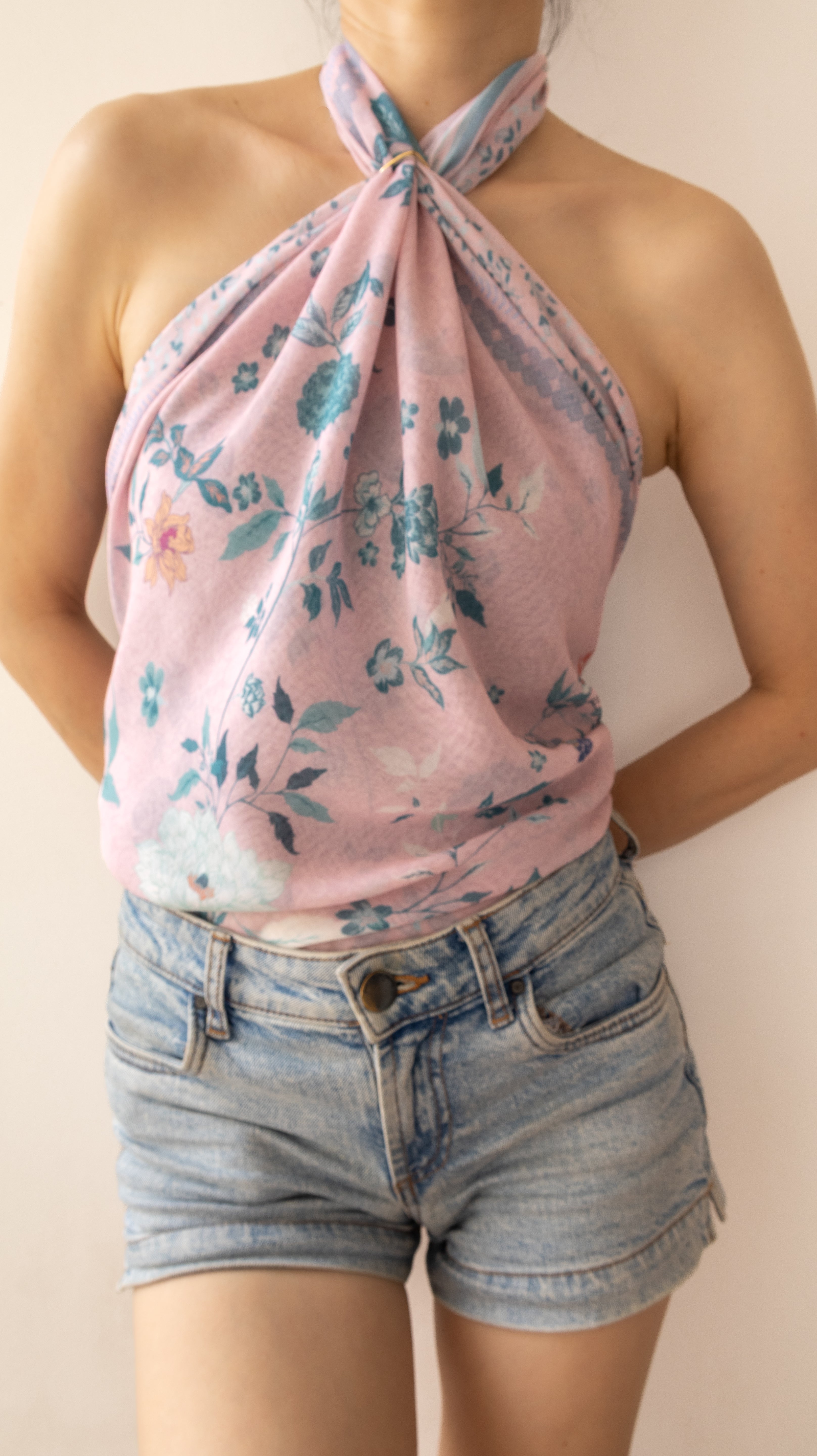 A woman wearing a pink floral halter top with blue and teal botanical print, paired with light-wash denim shorts, against a plain beige background.