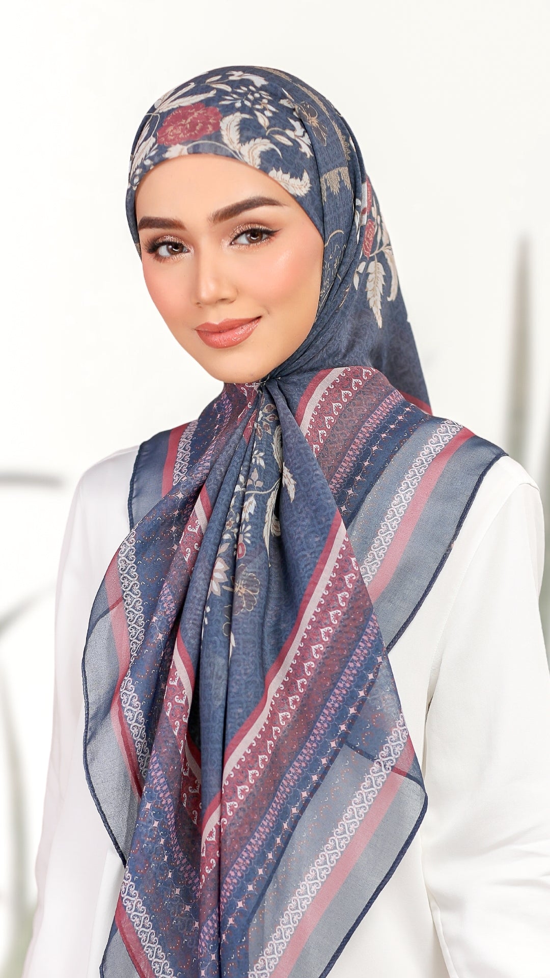 A woman wearing a patterned blue hijab with red and white geometric stripes, paired with a white top, set against a bright, softly blurred background.