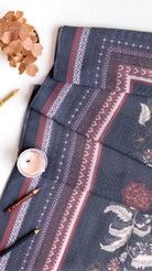 A folded dark slate blue fabric with intricate geometric patterns and floral motifs in dusty pink, burgundy, and cream, styled on a white surface with decorative props.