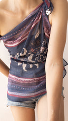 A close-up of a model wearing a navy blue one-shoulder wrap top with a floral print and patterned borders, paired with denim shorts.