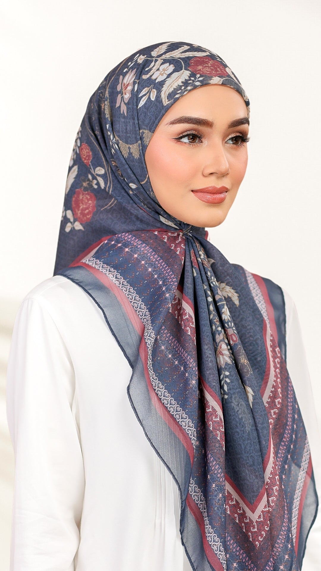 A woman wearing a navy blue patterned hijab with red and beige floral and geometric designs, paired with a white top, set against a plain white background.
