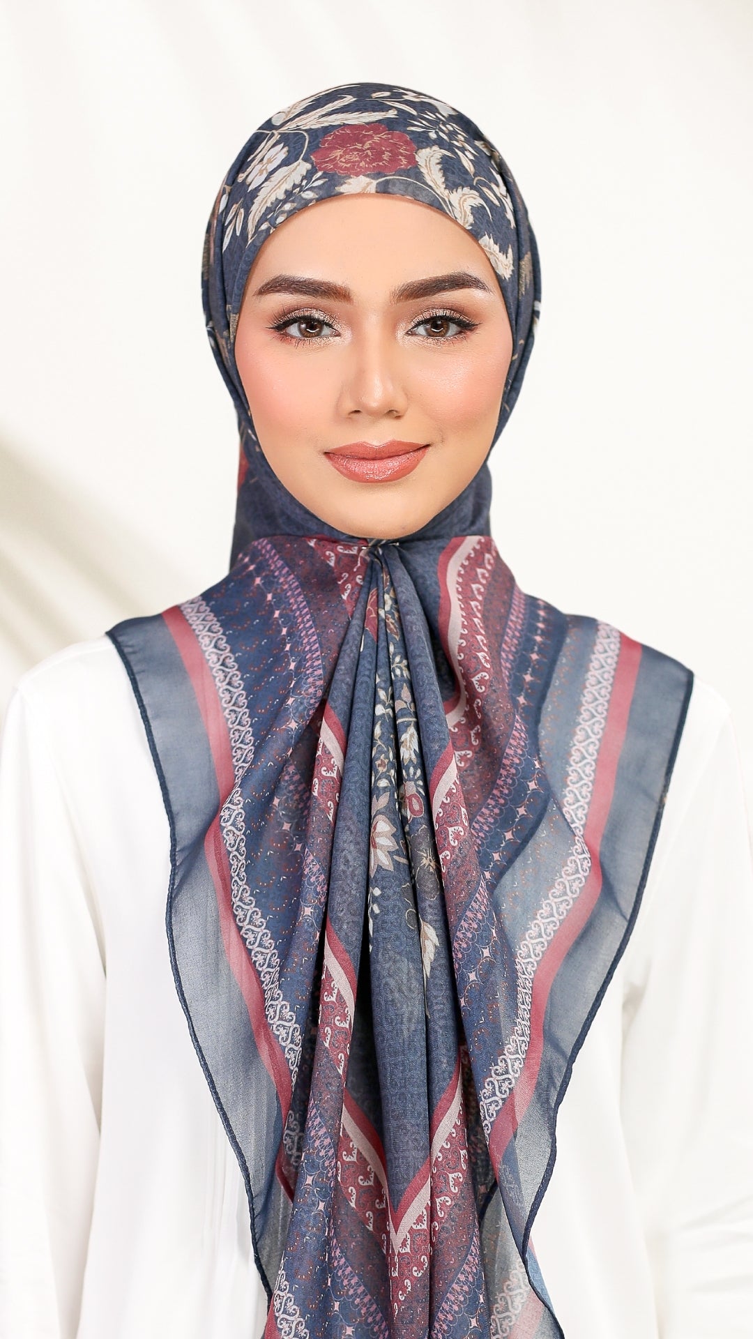 A woman wearing a patterned hijab in navy blue, dusty pink, and beige, paired with a white top against a soft white background.