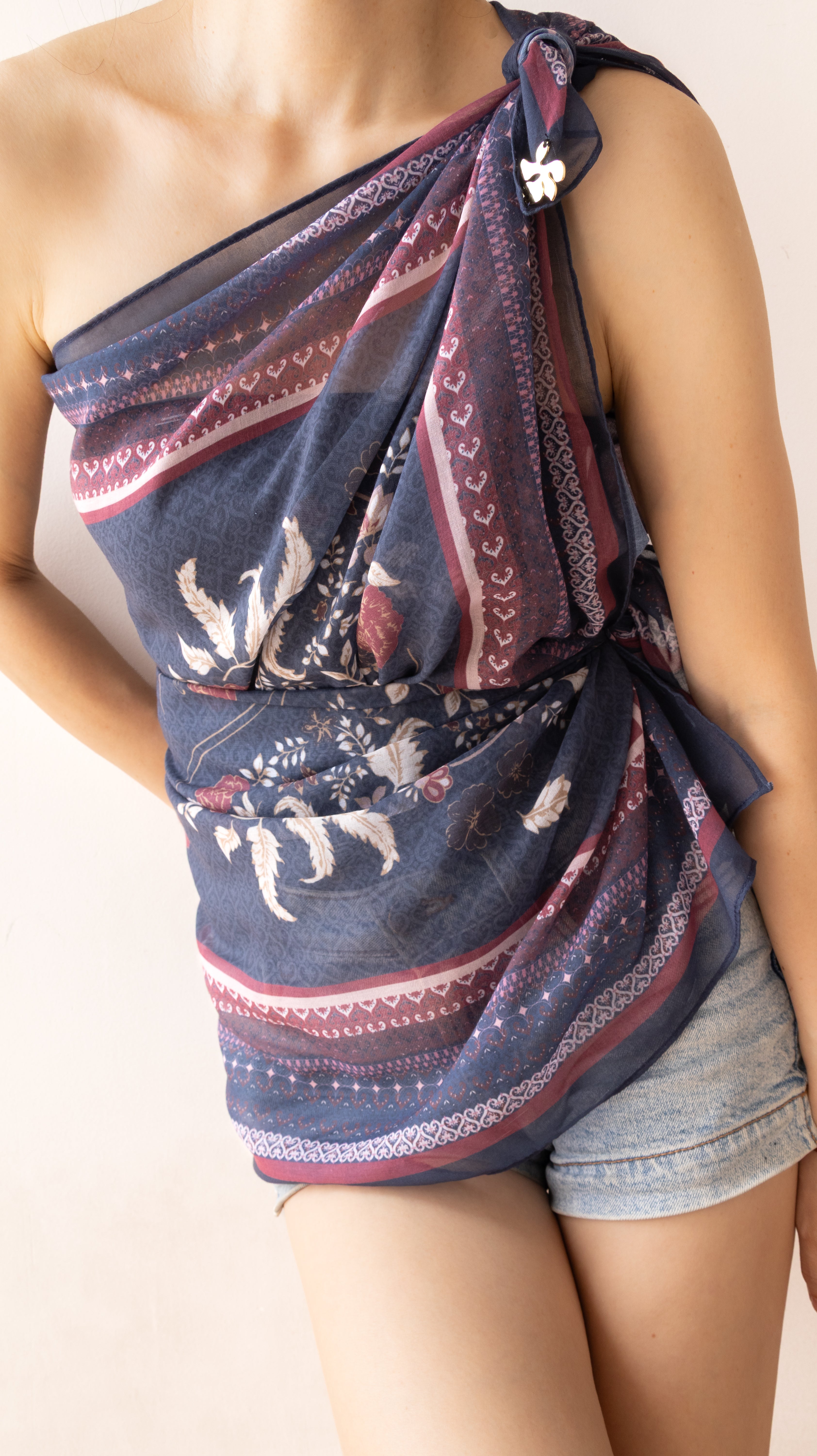 A woman wearing a draped, one-shoulder wrap top made from a navy blue patterned scarf with red and white accents, paired with denim shorts.