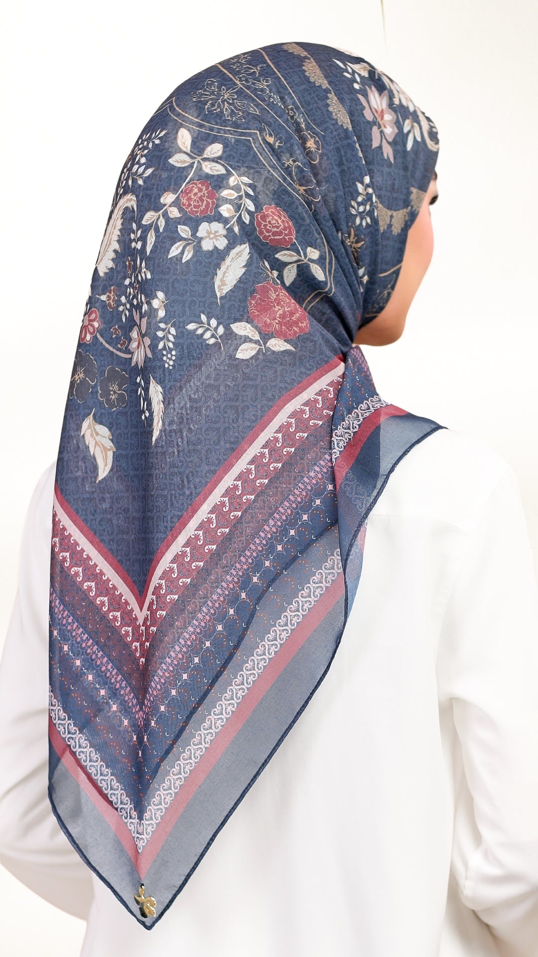A woman shown from the back wearing a navy blue patterned hijab with floral motifs and geometric borders, paired with a white top against a plain white background.