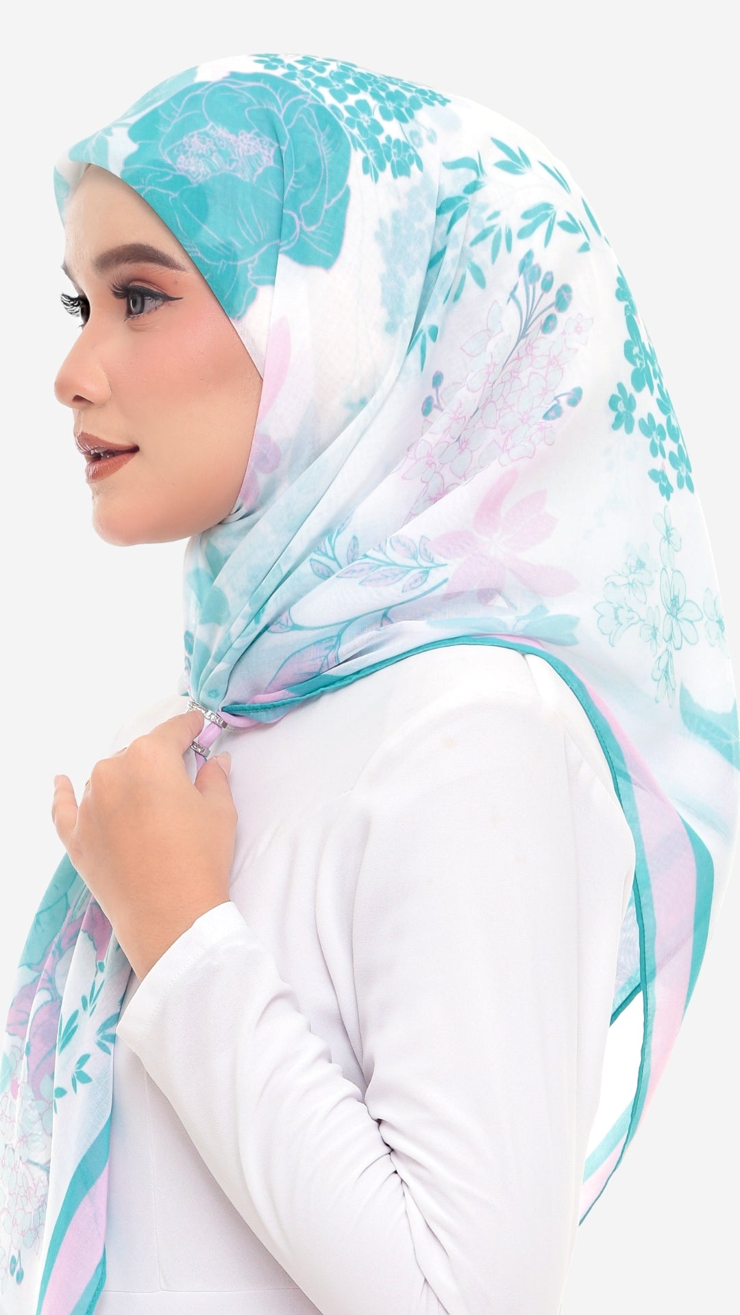 A woman in profile wearing a white hijab with a light blue and pink floral pattern, paired with a white long-sleeve top, against a plain white background.