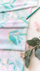 A styled flat-lay of light blue fabric with a pastel floral and leaf print, arranged on a cream surface with a green pencil, pink thread, and green leaves with small pink flowers.