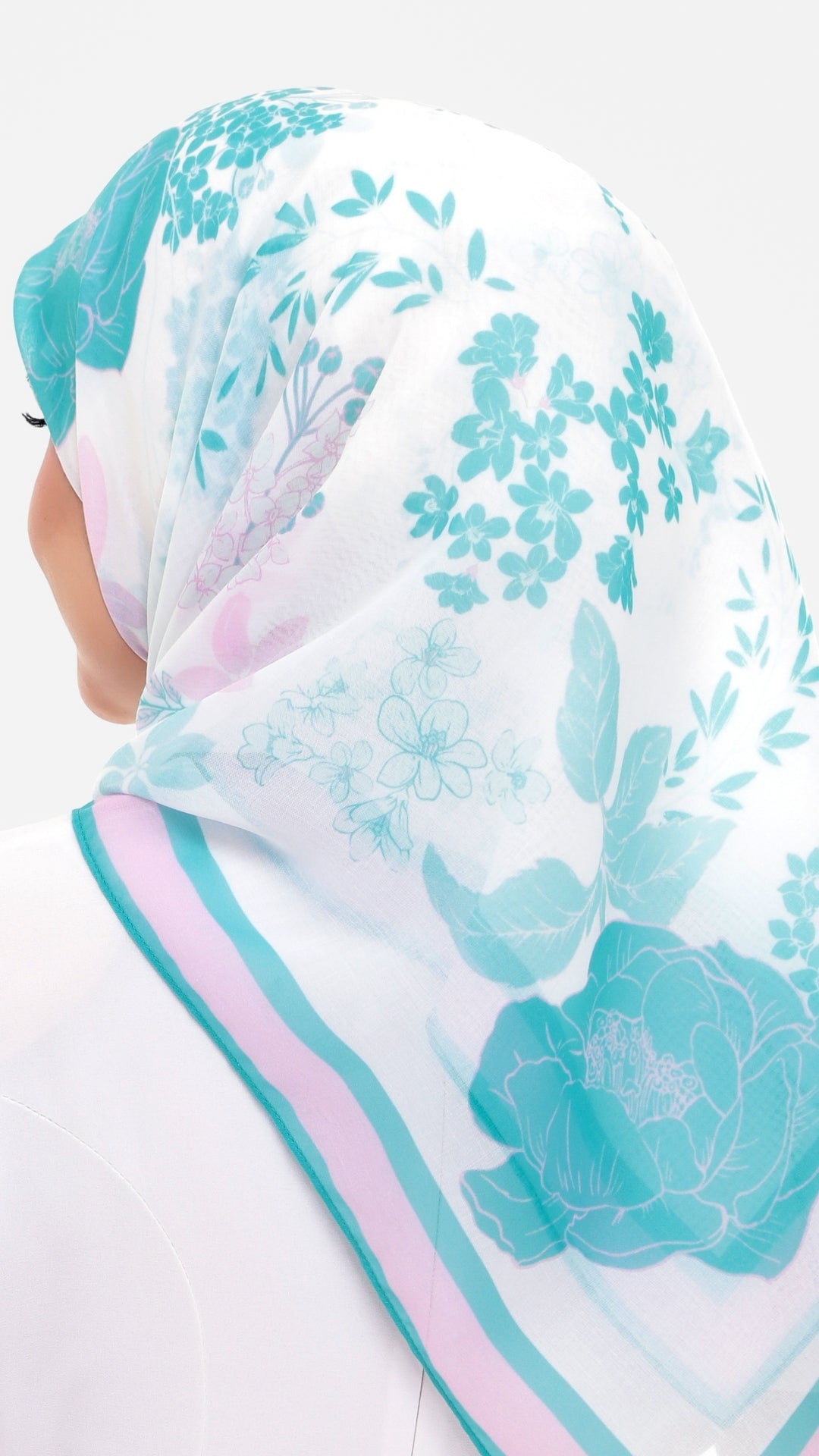 A close-up of a person wearing a white hijab with a teal floral pattern and pink striped border, shown from the back against a plain white background.