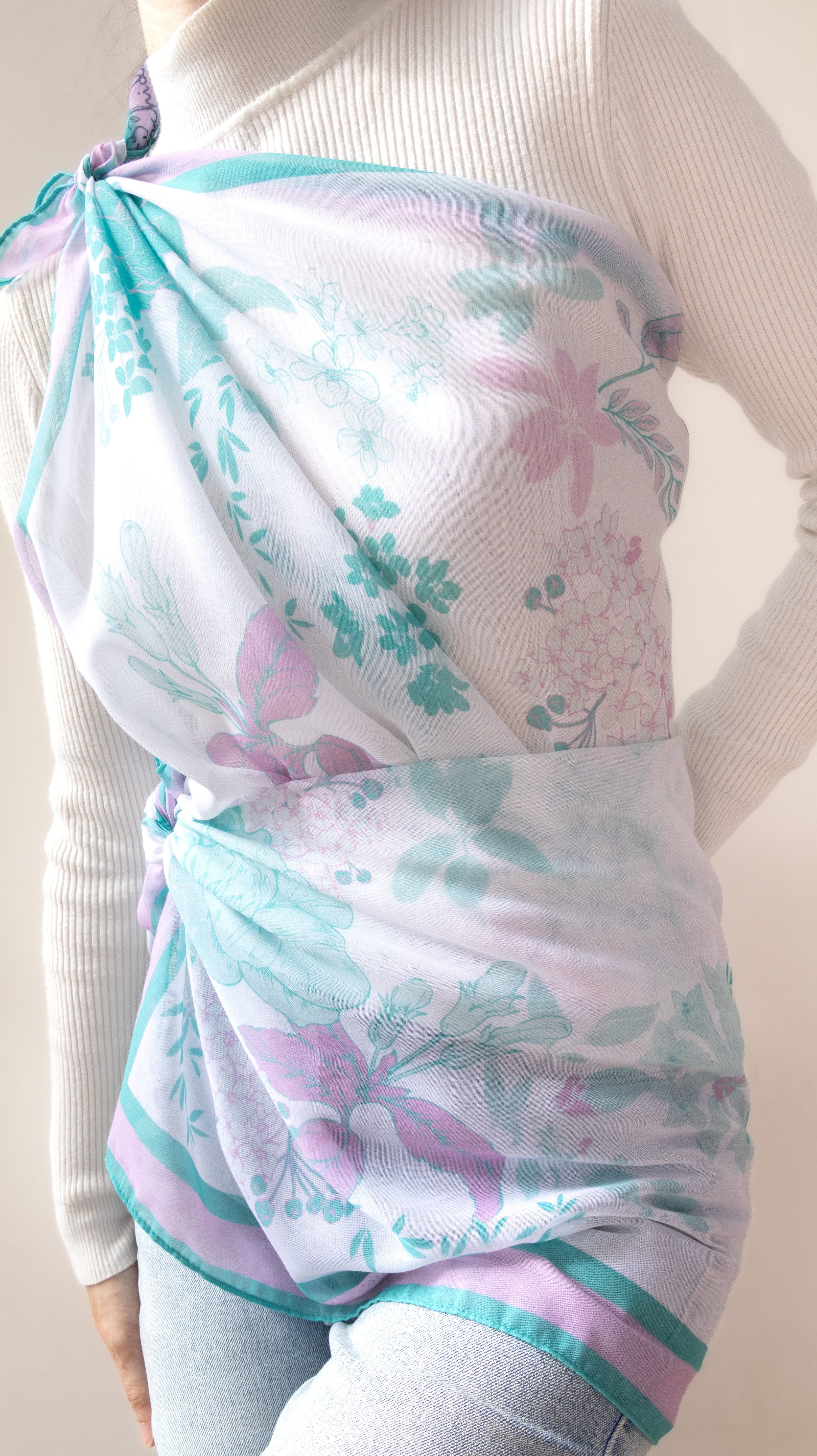 A white sheer wrap or scarf with a pastel floral print in mint green, lavender, and pink, styled as a shoulder drape over a white ribbed turtleneck sweater.