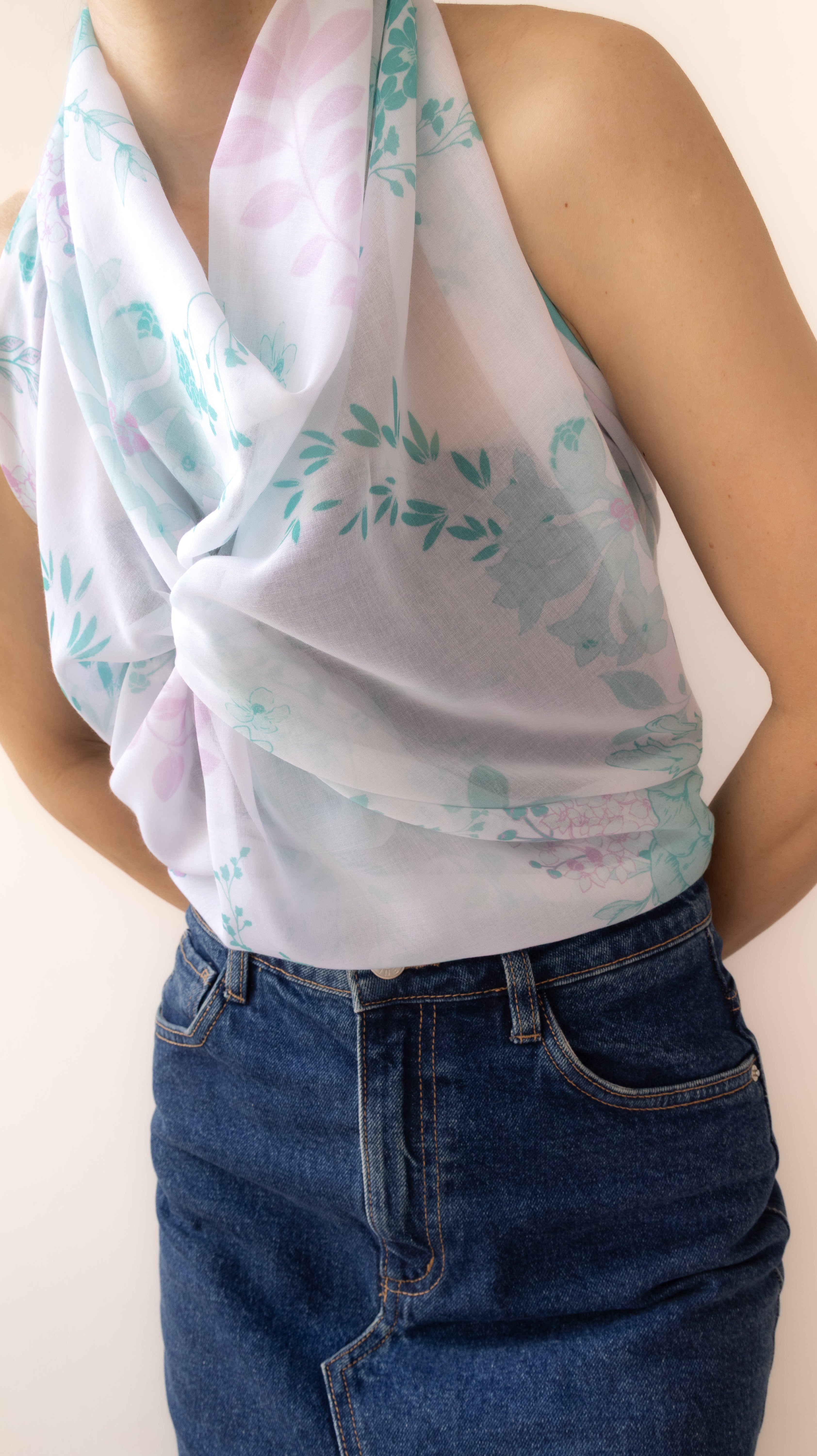 A close-up of a person wearing a sheer white halter-style scarf with a pastel floral print in pink and mint green, paired with dark blue jeans against a plain light background.