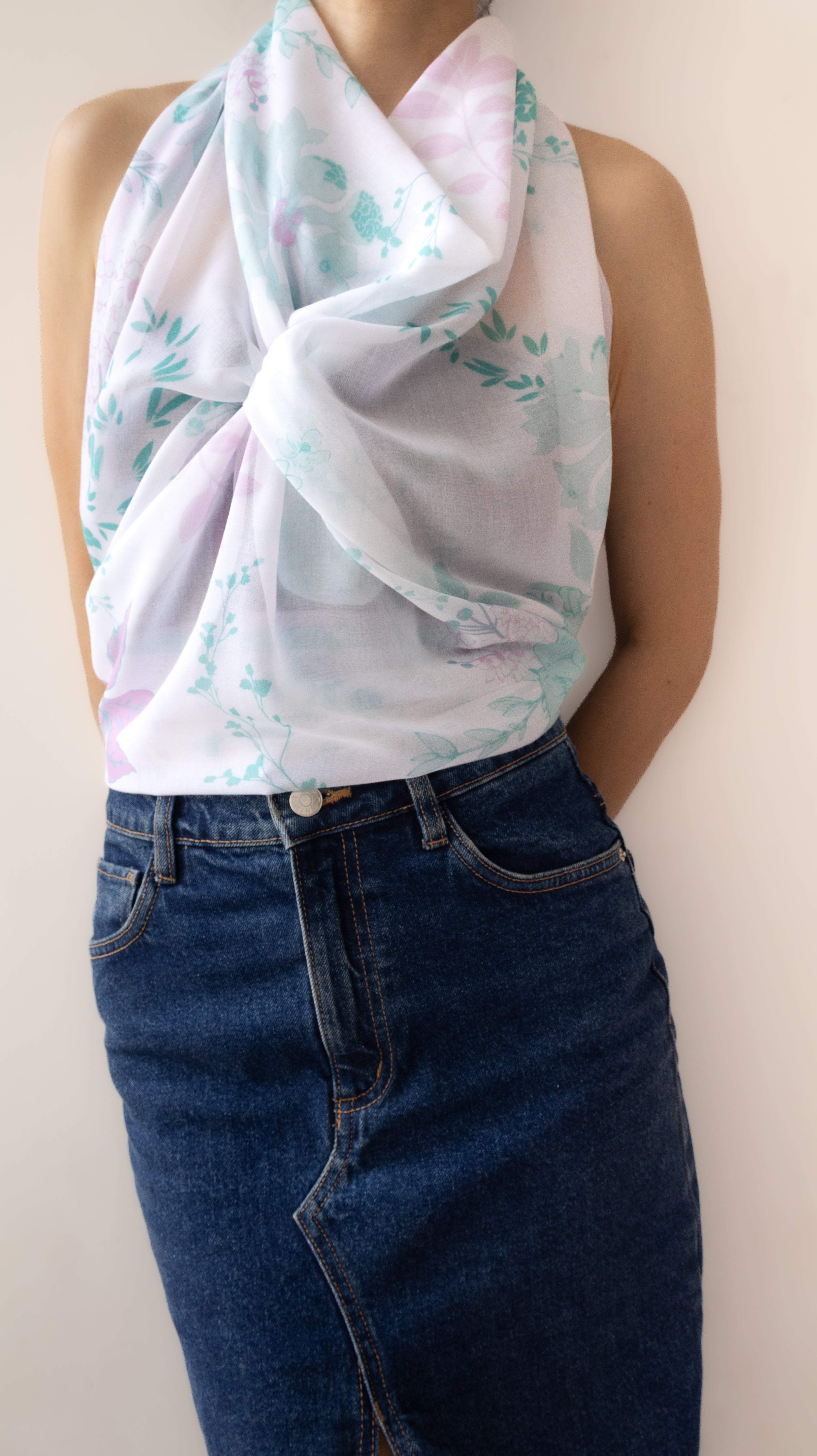 A sleeveless white scarf with a light blue and pink watercolor floral pattern, styled as a knot over the shoulders of a model wearing blue denim jeans.