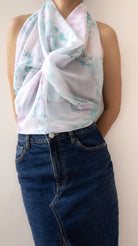 A sleeveless white scarf with a light blue and pink watercolor floral pattern, styled as a knot over the shoulders of a model wearing blue denim jeans.