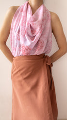 A person wearing a draped light pink scarf with a darker pink floral pattern over a sleeveless top and a rust-brown wrap skirt, shown against a plain light beige wall.