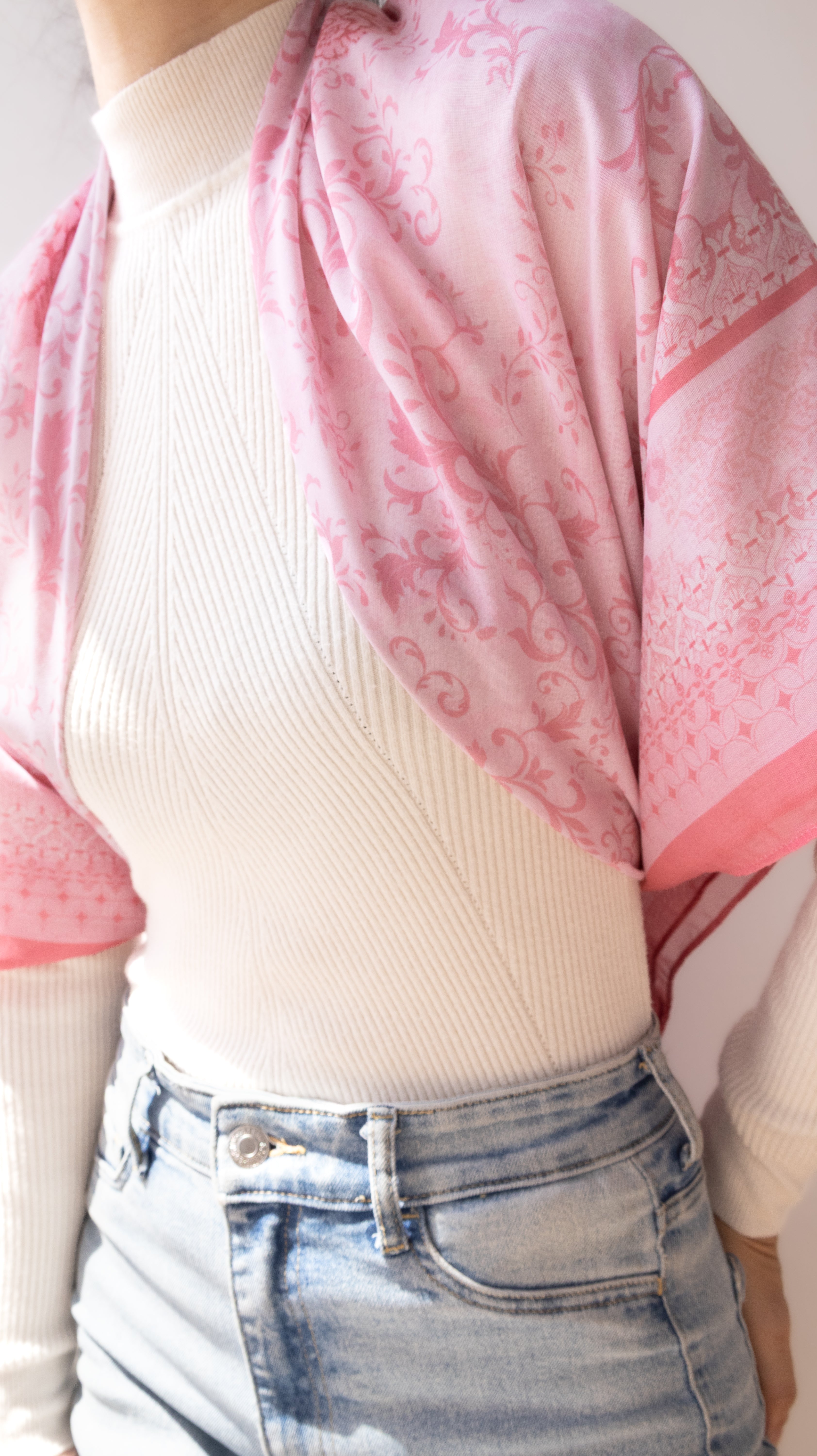 A close-up of a person wearing a light pink patterned shawl draped over a cream ribbed turtleneck sweater and light-wash blue jeans.