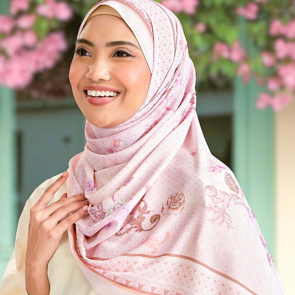 Woman wearing a pink hijab with floral patterns against a blurred background of pink flowers and greenery.