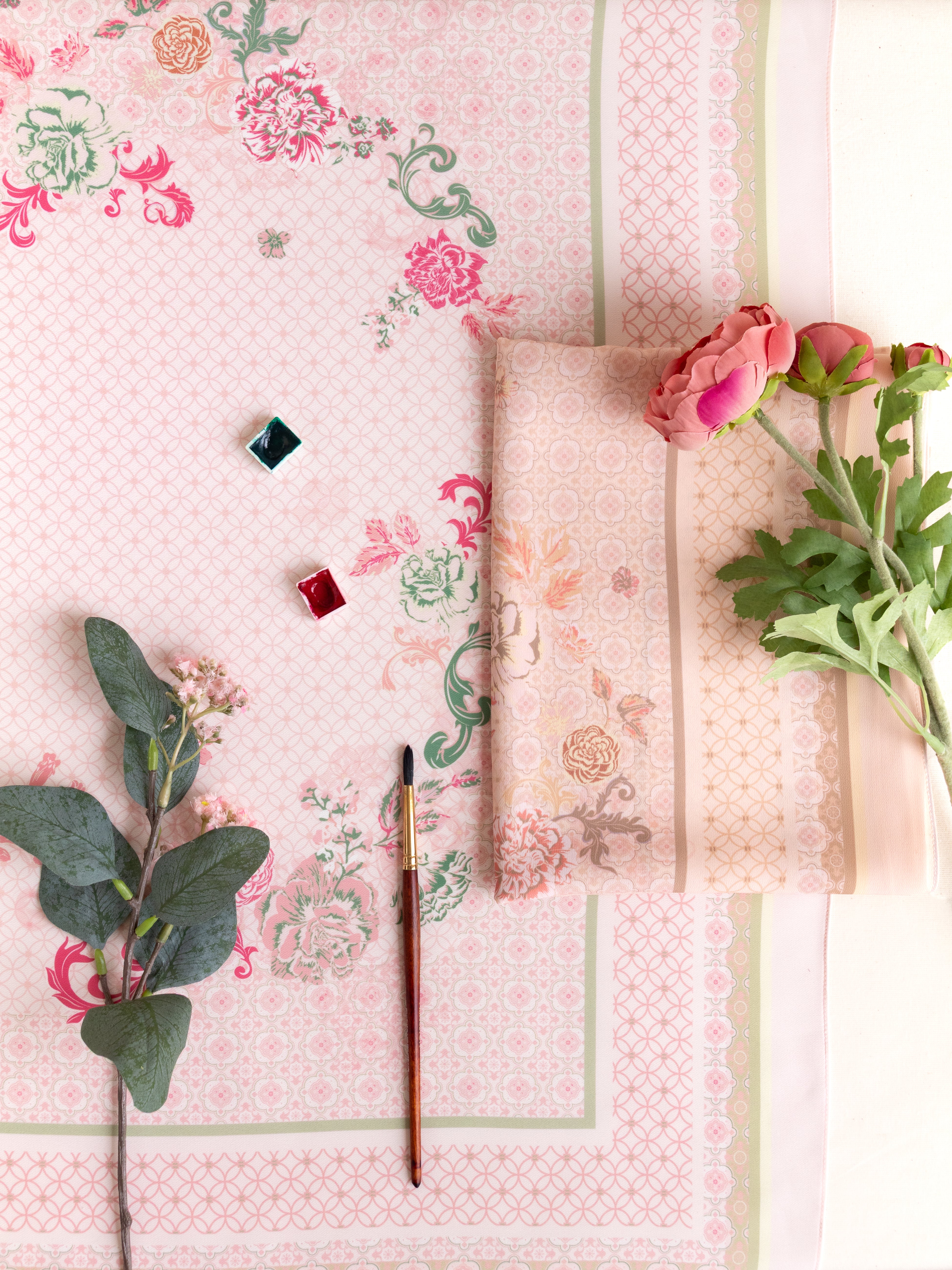 A styled flat-lay of a light pink floral fabric with a geometric border, featuring a folded matching fabric piece, a paintbrush, small paint cubes, and pink flowers on a white background.