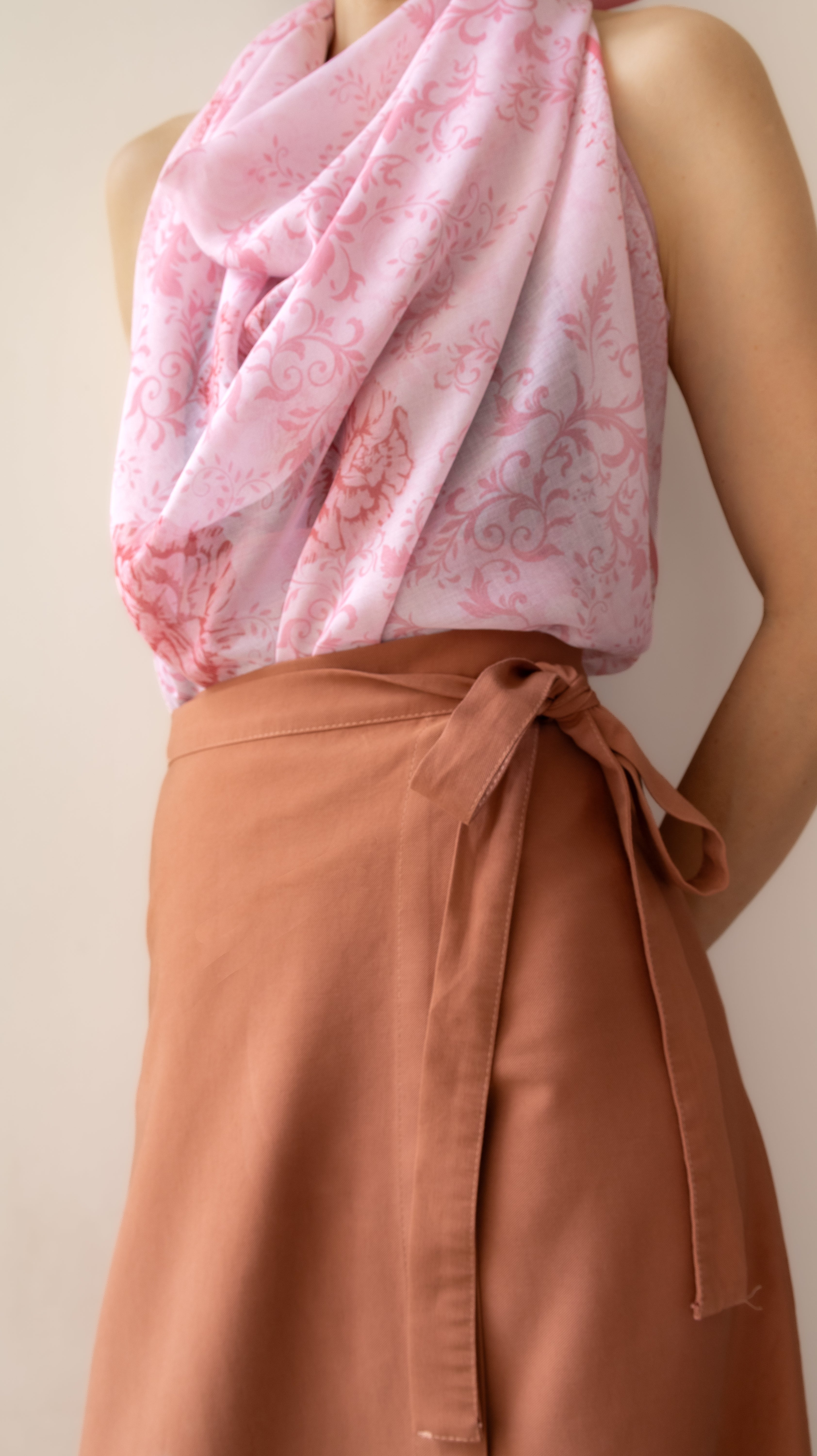 A close-up of a person wearing a draped light pink patterned top and a rust-brown wrap skirt tied at the waist, against a plain beige background.