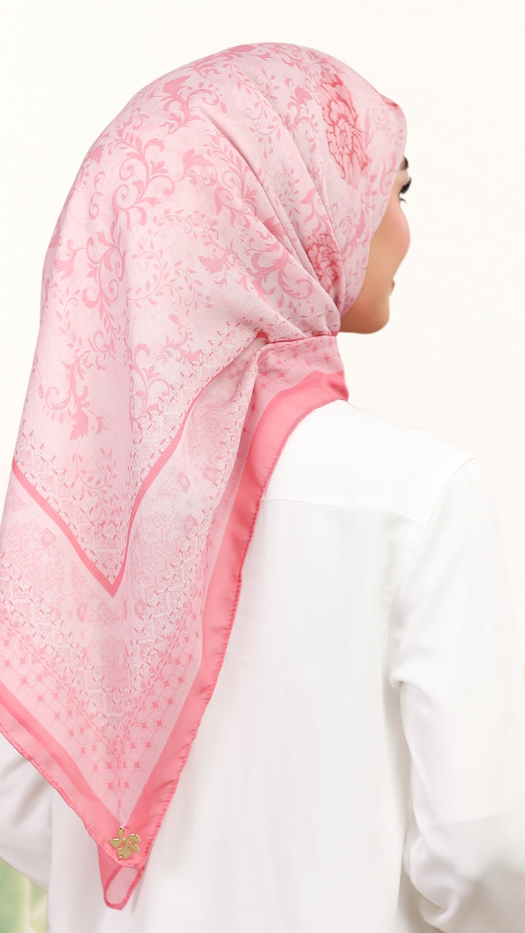 A woman shown from the back wearing a light pink patterned hijab and a white long-sleeve top against a plain white background.