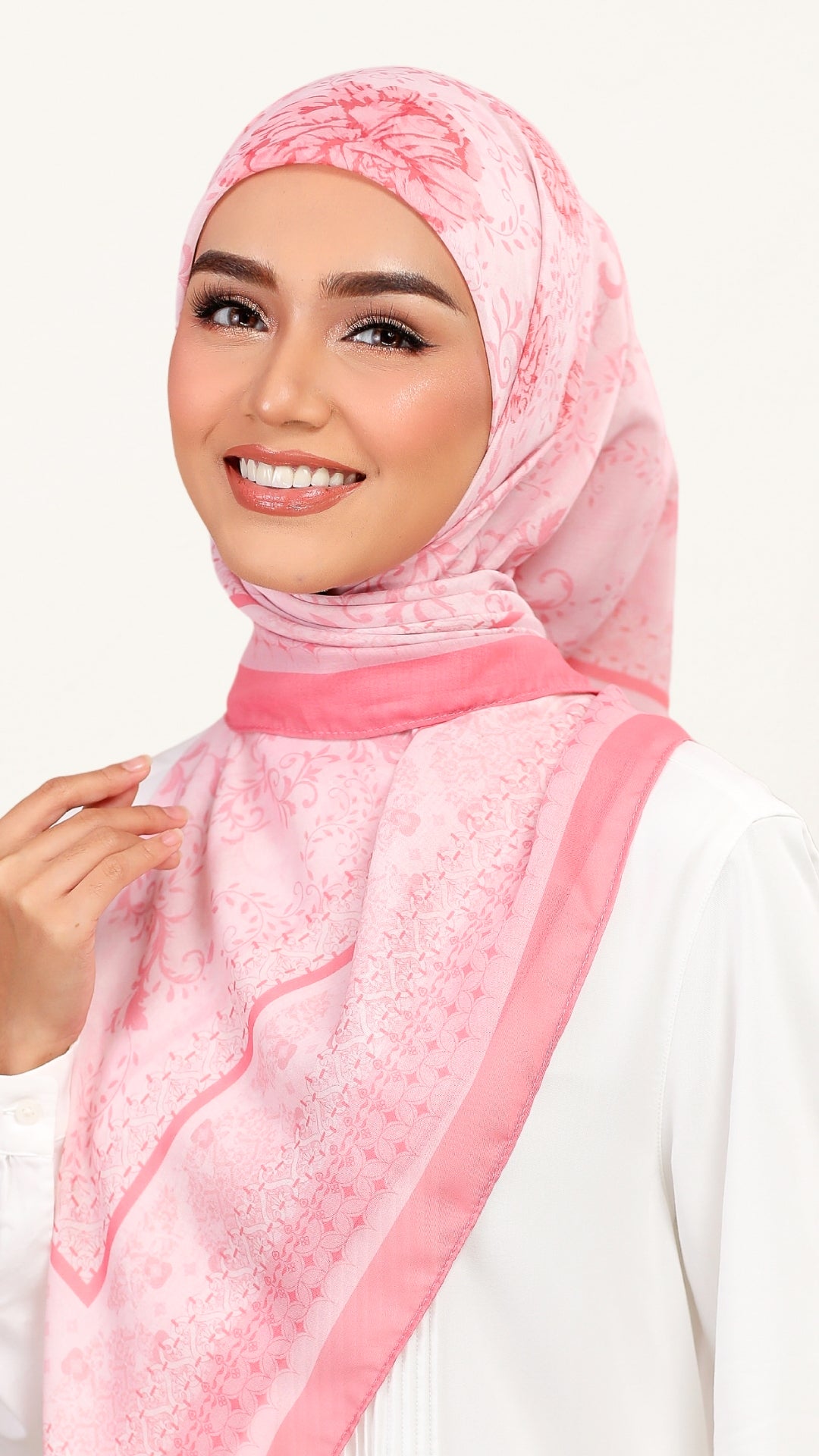 A smiling woman wearing a light pink patterned hijab with a darker pink border, paired with a white top, set against a plain white background.