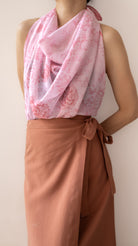 A model wearing a light pink draped scarf with a darker pink floral pattern, styled over a sleeveless top and a rust-brown wrap skirt against a plain beige background.