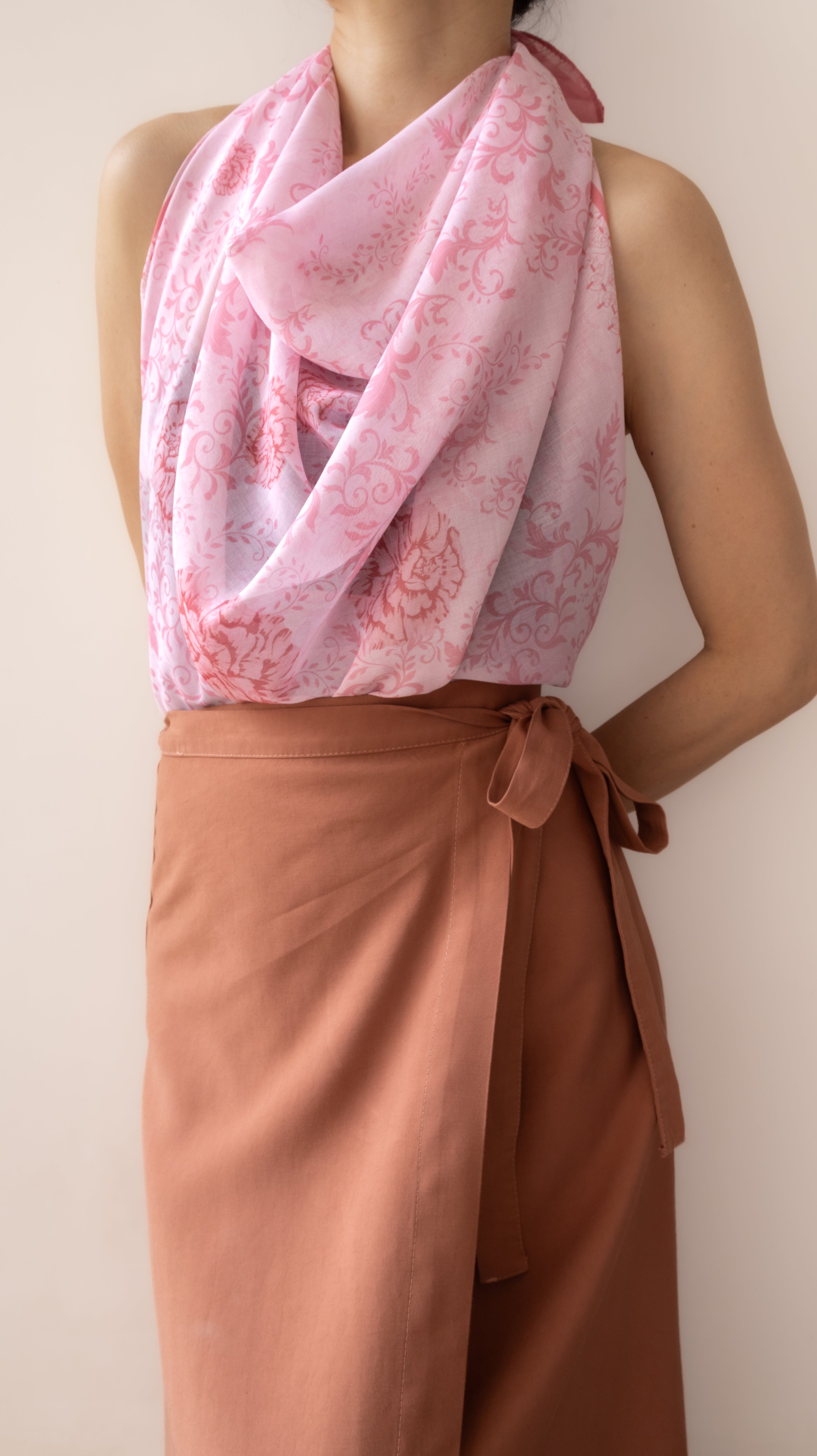 A model wearing a light pink draped scarf with a darker pink floral pattern, styled over a sleeveless top and a rust-brown wrap skirt against a plain beige background.