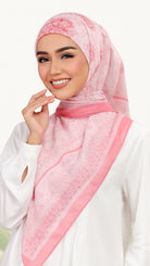 A woman wearing a light pink patterned hijab with a darker pink border, paired with a white long-sleeve top, set against a clean white background.