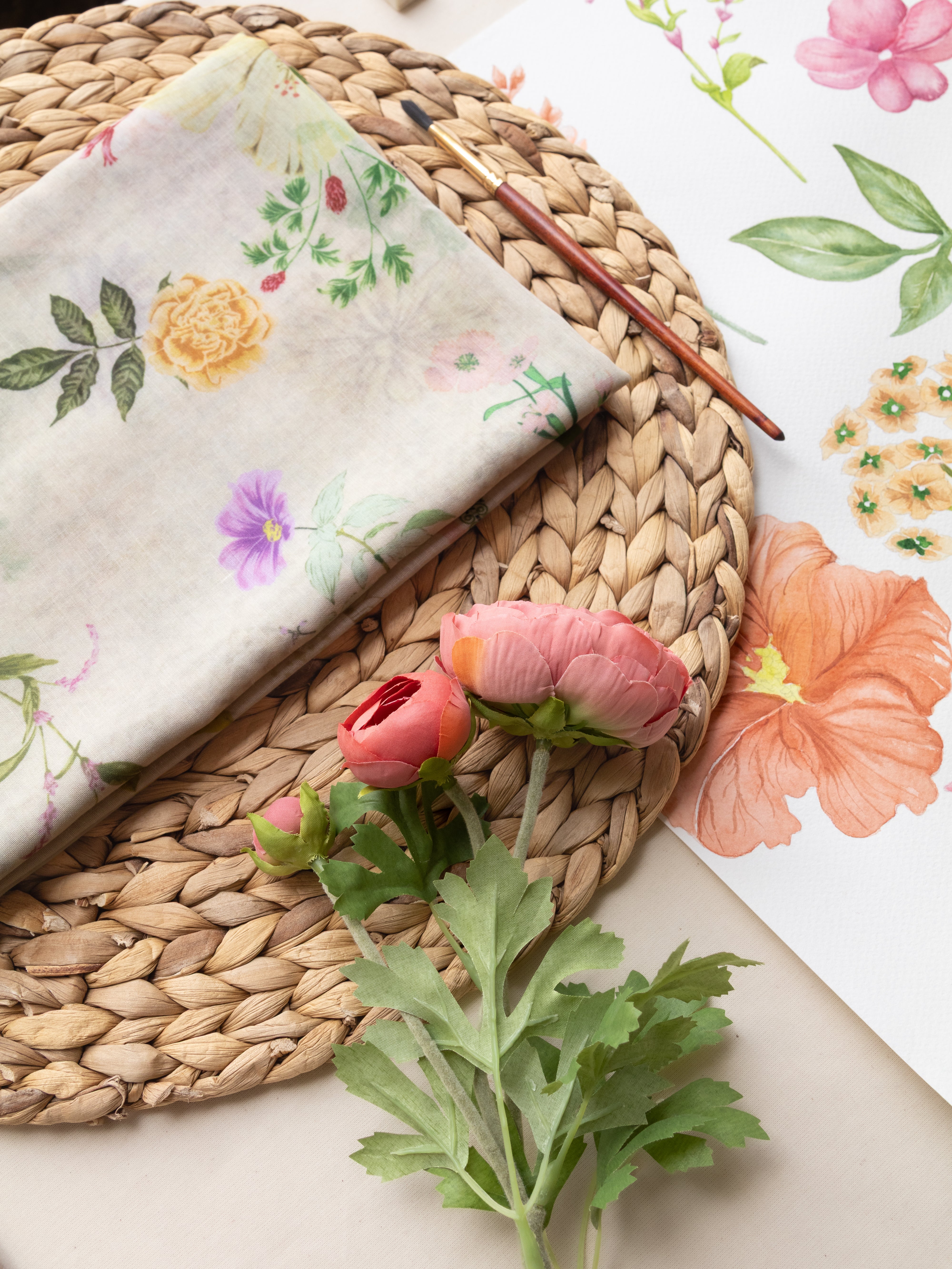 A folded cream fabric napkin with a delicate pastel floral print, styled on a round woven straw placemat alongside pink ranunculus flowers, a small paintbrush, and a sheet of floral paper.