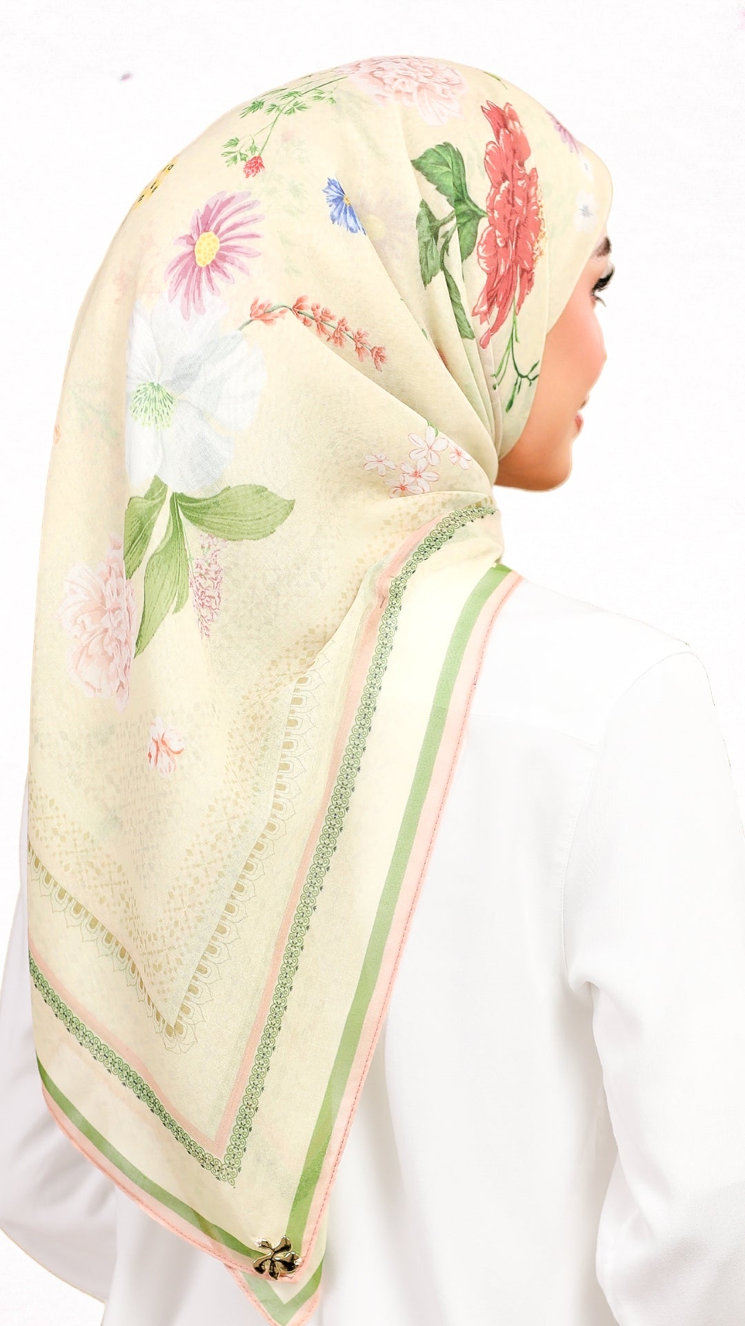 A woman shown from the back wearing a light beige hijab with a colorful floral print in pink, green, and white, paired with a white top against a plain white background.