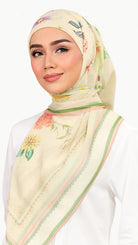 A woman wearing a light beige floral hijab with green and pink border stripes, styled over a white top against a plain white background.