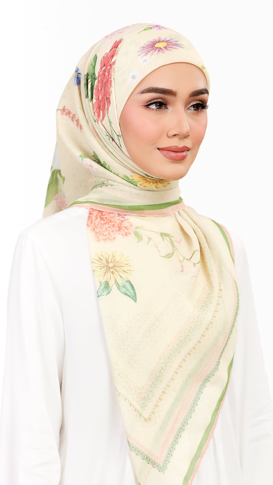 A woman wearing a cream-colored floral hijab with pink, green, and yellow accents, paired with a white long-sleeve top against a plain white background.