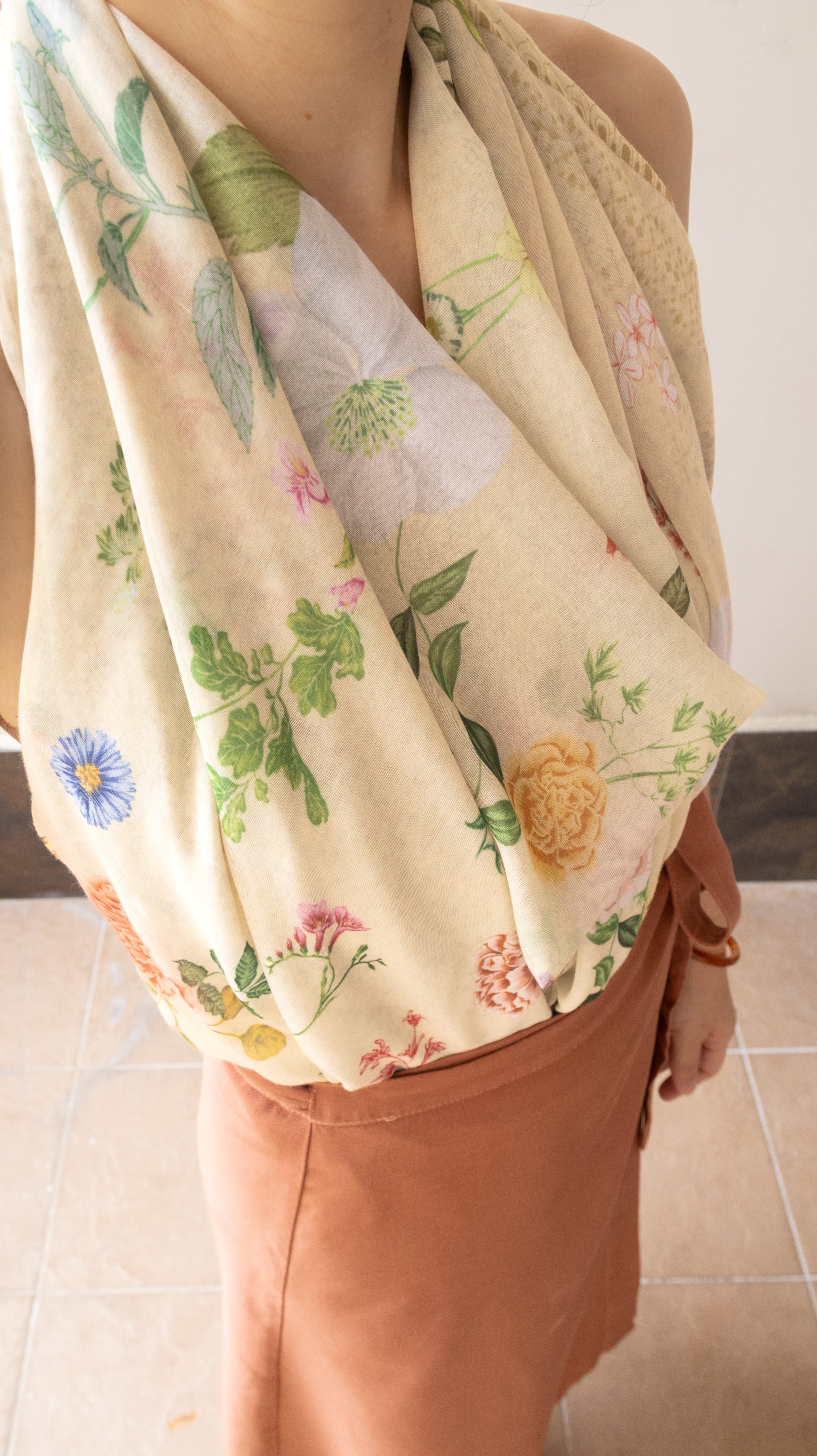 A close-up of a person wearing a cream-colored halter-style wrap or scarf with a colorful floral print, paired with a rust-orange skirt. The background shows a tiled floor and a plain wall.