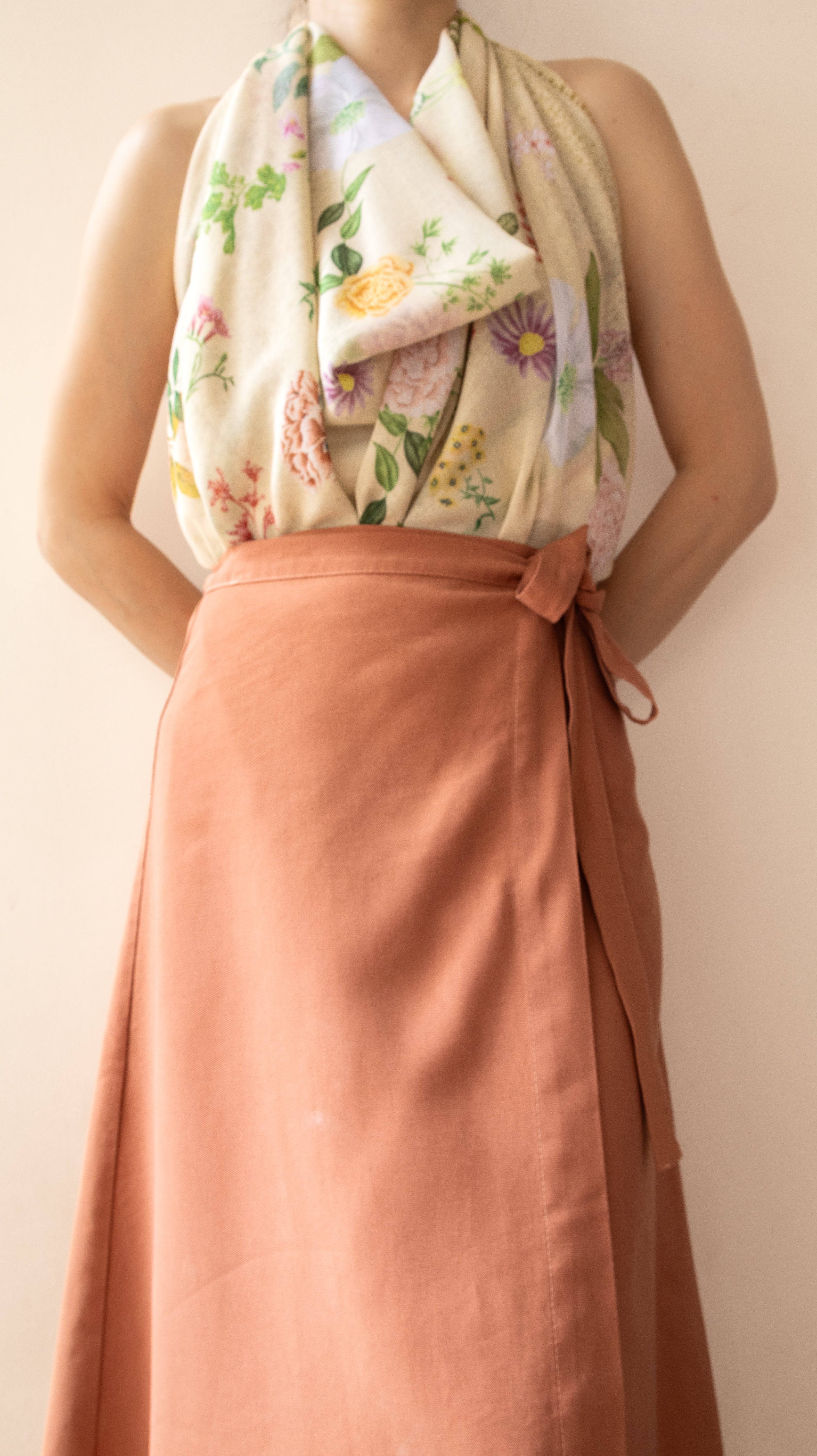 A person wearing a sleeveless rust-colored wrap skirt with a matching fabric tie at the waist, paired with a cream floral halter top against a plain beige background.