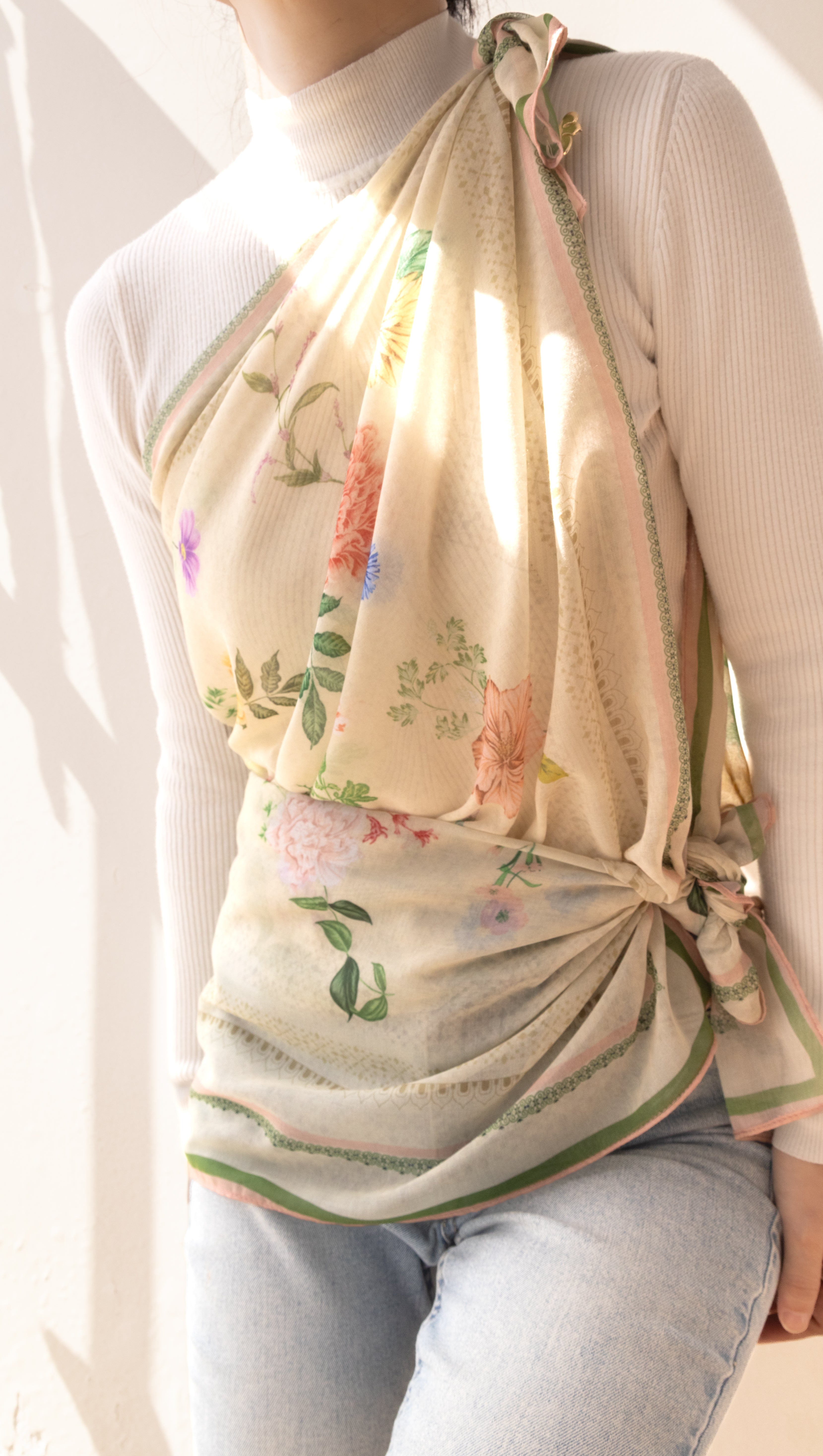 A cream-colored halter wrap scarf with a delicate pink, green, and orange floral print, styled on a model wearing a white ribbed turtleneck and light blue jeans.
