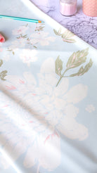 A close-up of a light blue floral fabric or wallpaper roll with large pale pink and white flowers, olive green leaves, and soft blush accents. A teal pencil and pink cosmetic items are partially visible on a lavender surface.