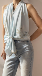 A sleeveless light blue halter-style top with a draped neckline and soft floral embroidery, tied at the waist and worn with light-wash jeans against a plain beige studio background.