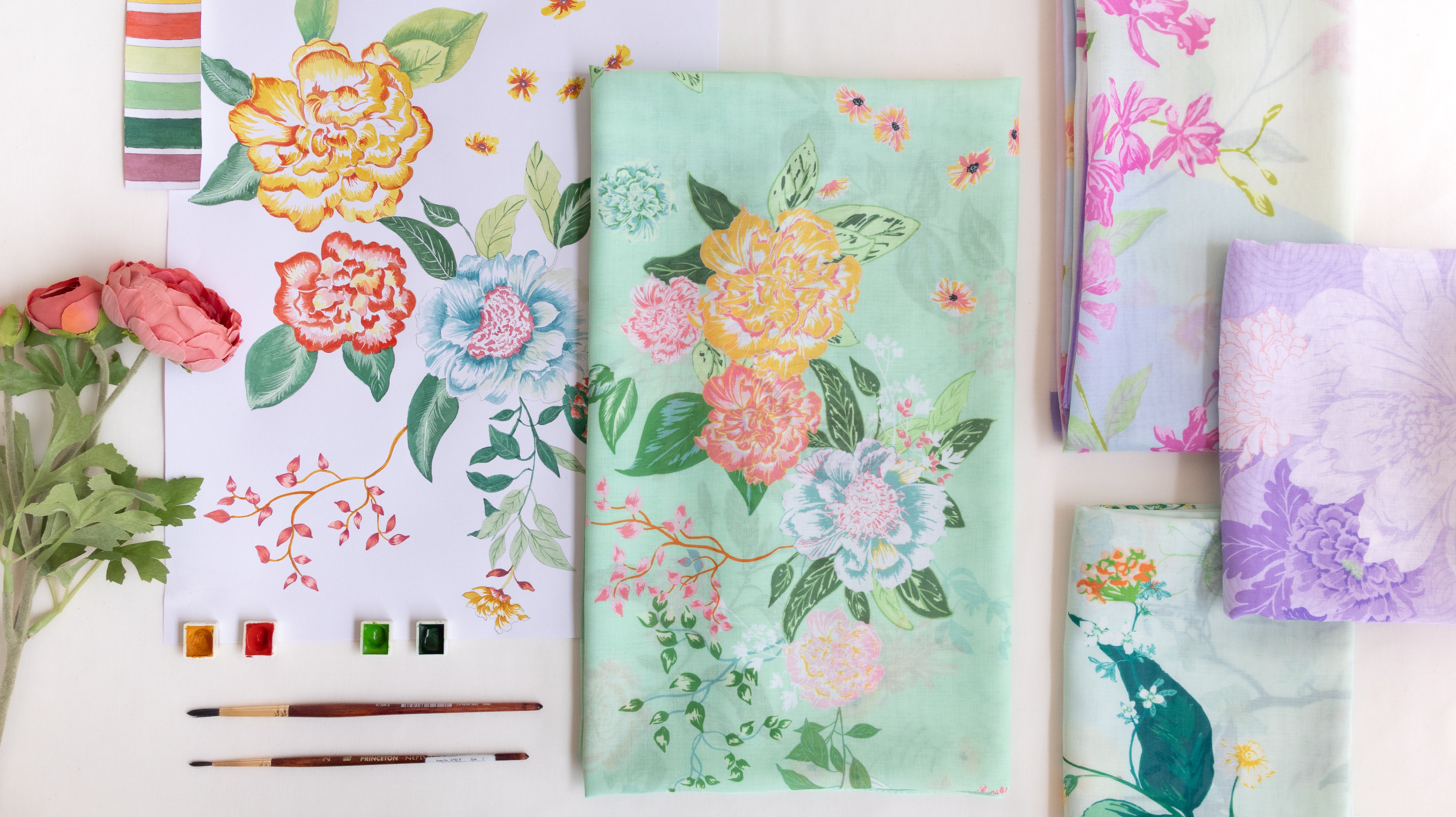 A styled flat-lay of floral fabric or paper sheets with watercolor-style flowers and leaves. The central item is a mint green sheet with large peach, pink, blue, and yellow flowers and green leaves.