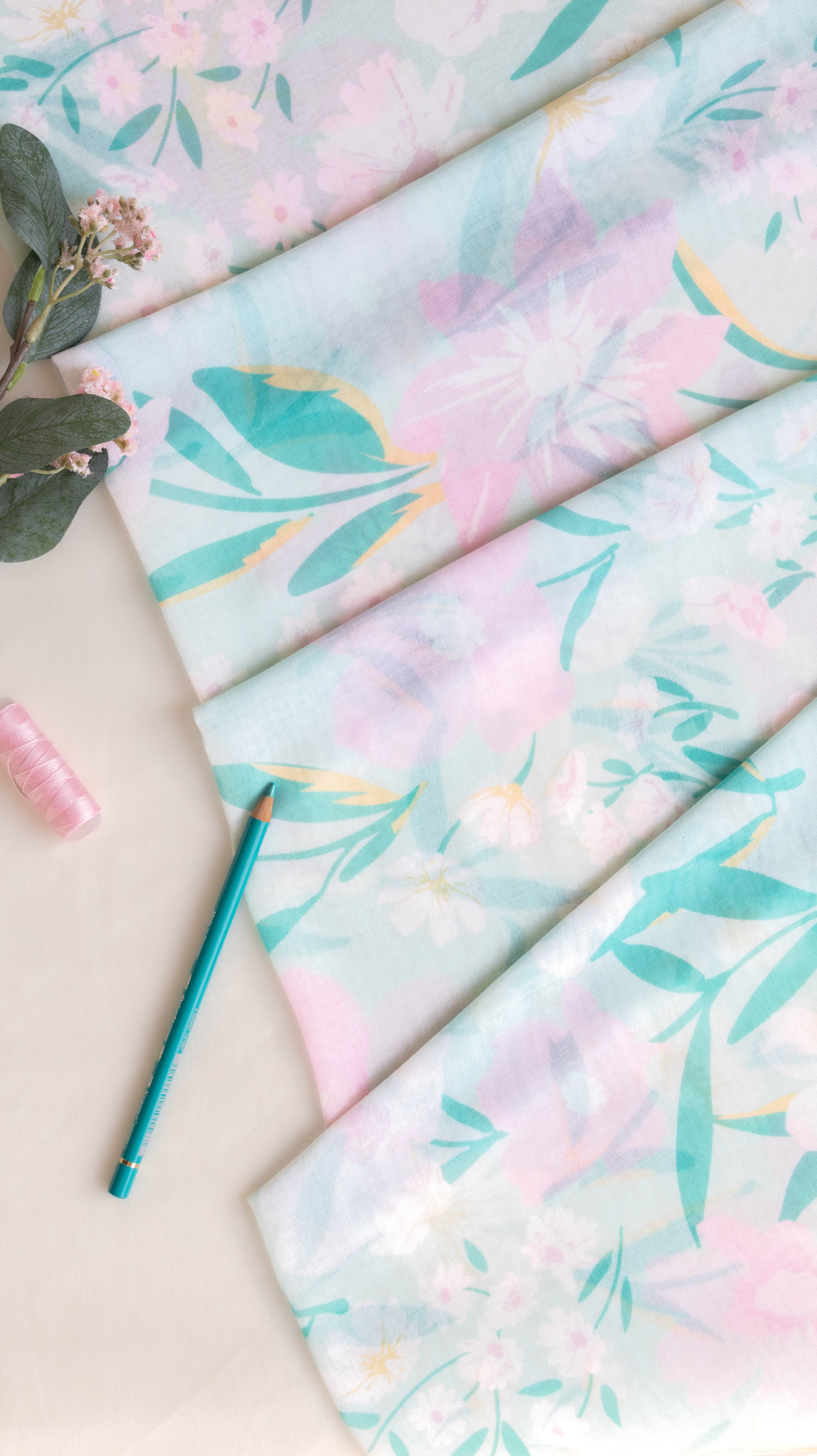 A folded piece of lightweight fabric with a pastel floral print in pink, mint green, and lavender, styled on a light beige surface with a teal pencil, a pink spool of thread, and eucalyptus leaves.