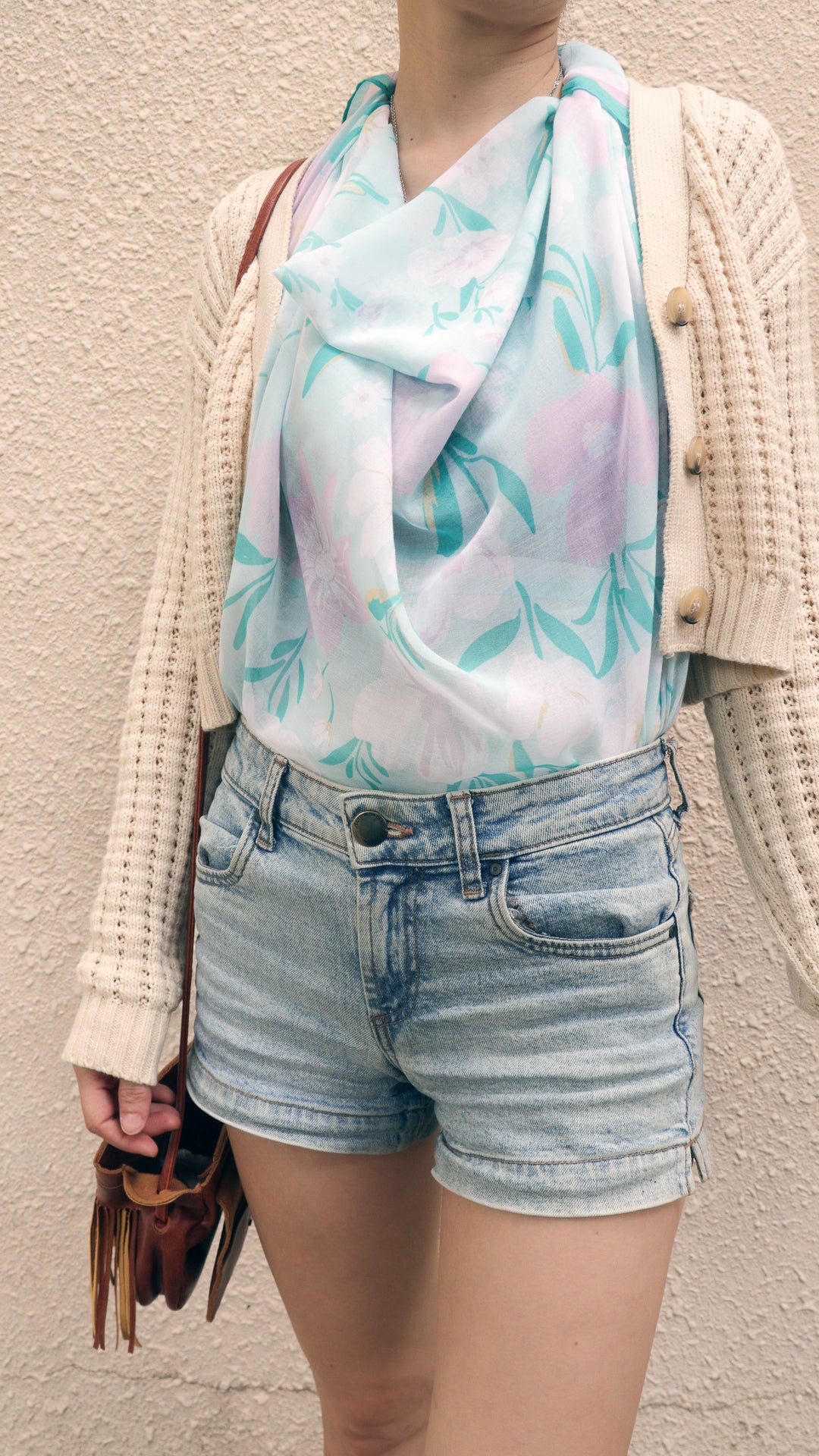 A person wearing a light blue and lavender floral print scarf, a cream knit cardigan, light-wash denim shorts, and holding a brown leather fringe bag against a textured beige wall.