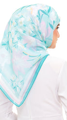 A woman wearing a light aqua hijab with a pastel floral print in pink, white, and green, paired with a white top against a plain white background.