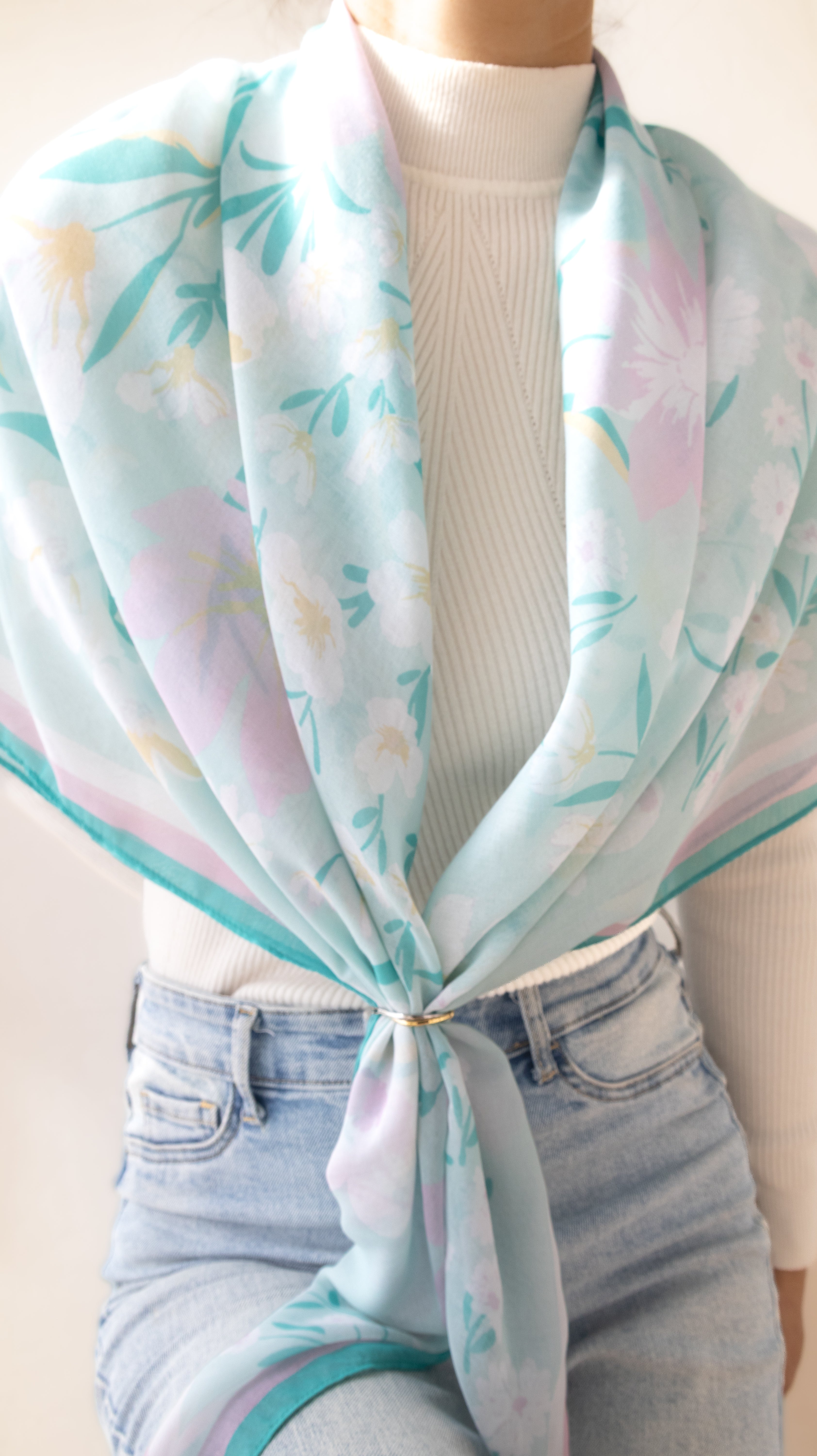A lightweight square scarf with a pastel floral print in mint green, lavender, and soft pink, styled tied around the waist of a model wearing a white ribbed turtleneck and light blue jeans.