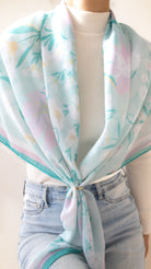 A lightweight square scarf with a pastel floral print in mint green, lavender, and soft pink, styled tied around the waist of a model wearing a white ribbed turtleneck and light blue jeans.