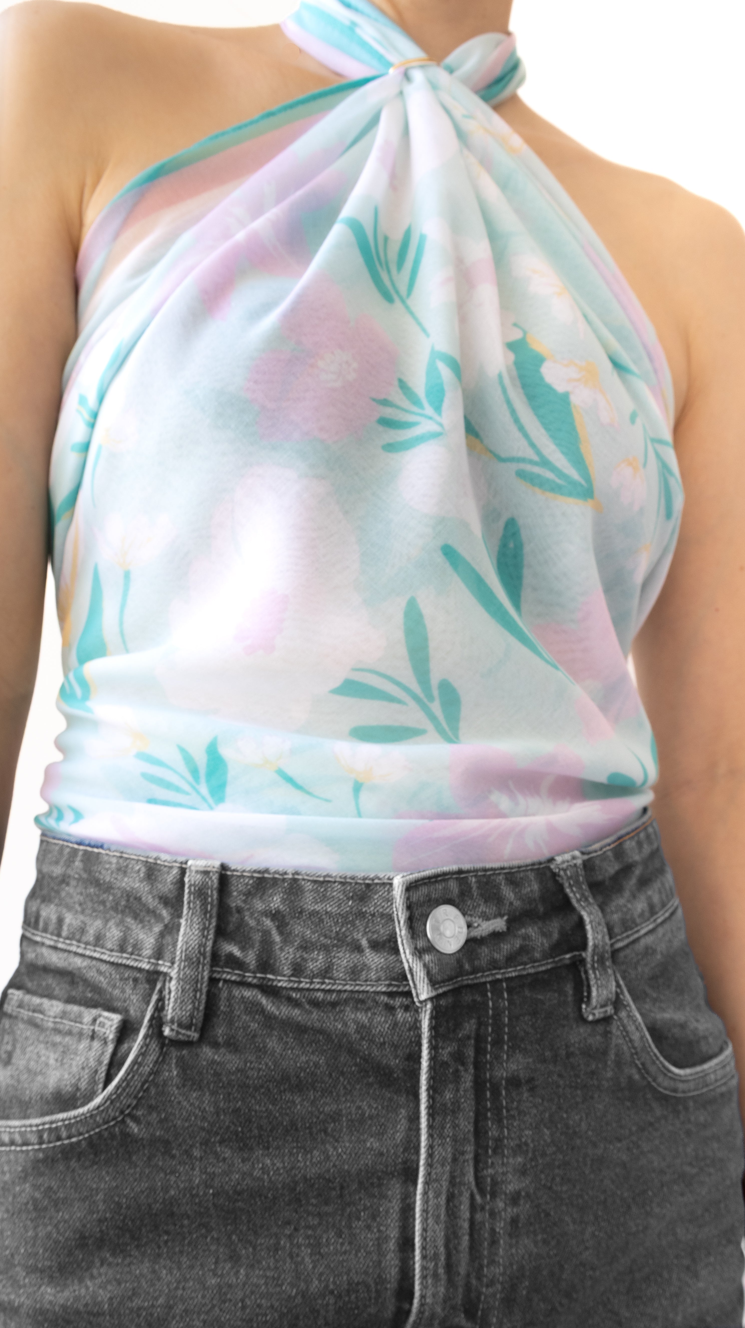 A close-up of a model wearing a sheer, halter-style top with a pastel floral print in pink, mint green, and light blue, paired with dark gray jeans.