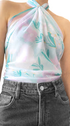 A close-up of a model wearing a sheer, halter-style top with a pastel floral print in pink, mint green, and light blue, paired with dark gray jeans.