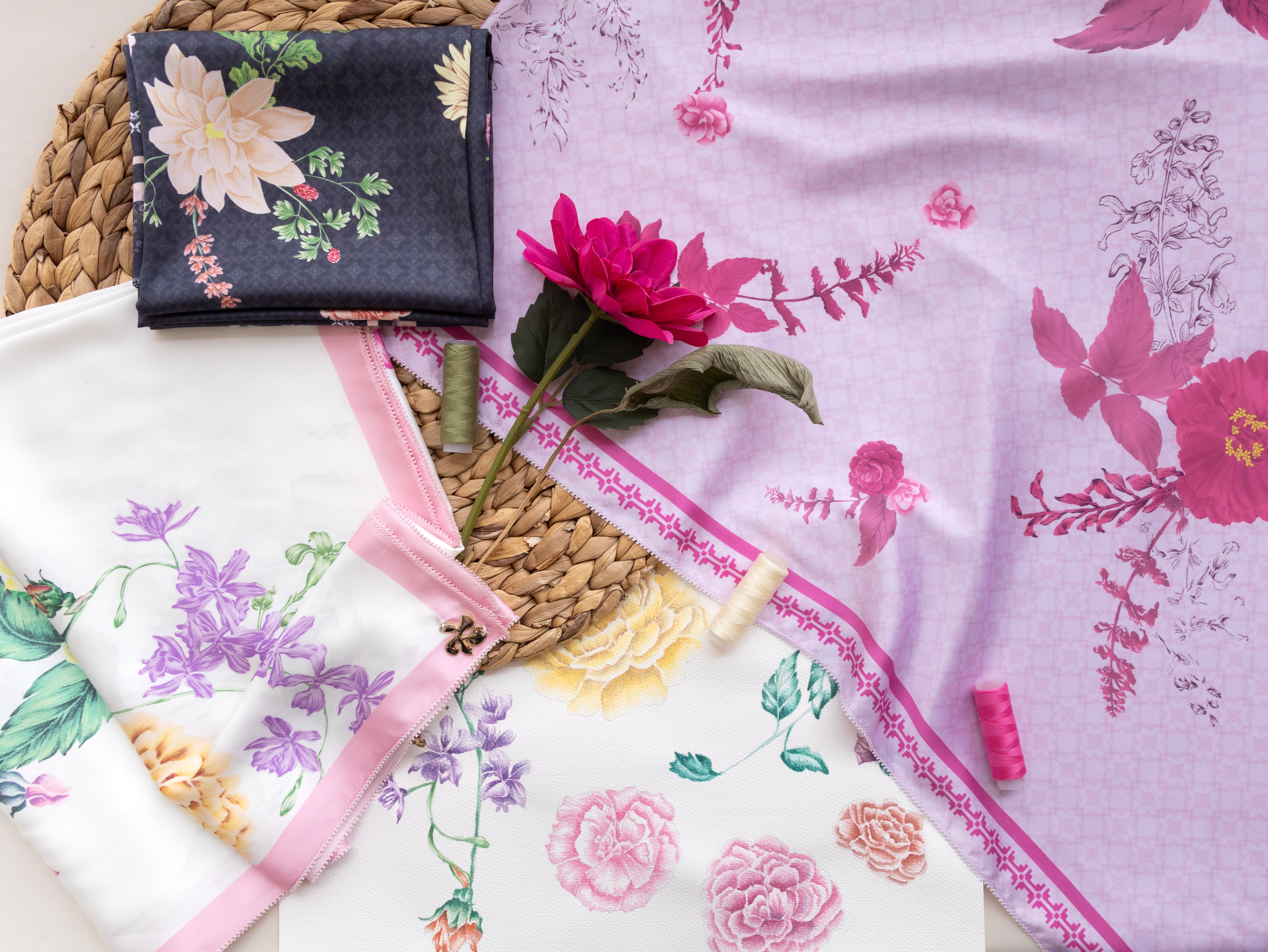 Three folded floral fabric pieces with pink borders are arranged on a woven mat. One is dark navy with large cream flowers, one is white with purple and yellow florals, and one is light lavender with pink floral print and border.