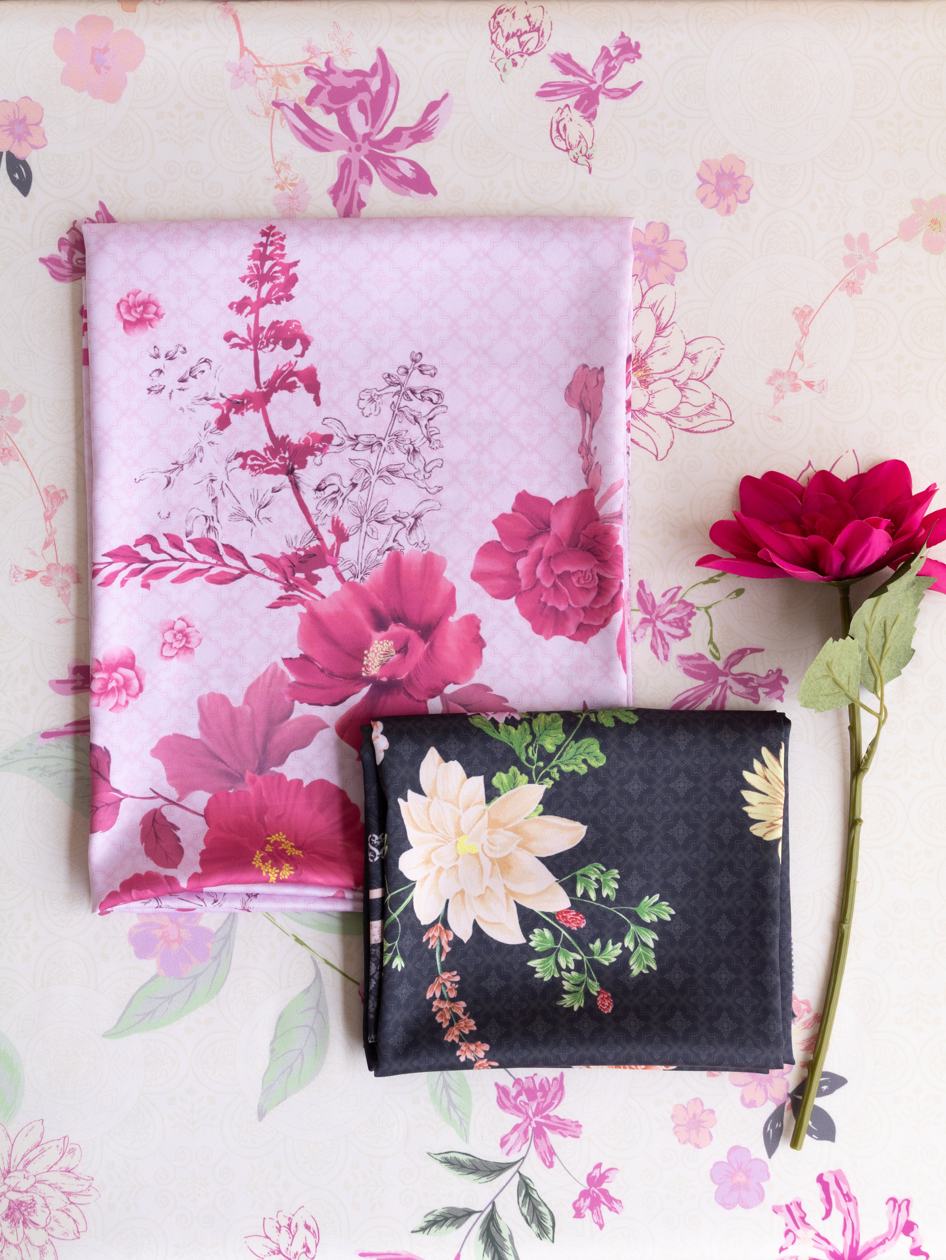 Two folded fabric pieces displayed on a floral background: a light pink fabric with large magenta and white flowers, and a dark charcoal fabric with cream flowers and green leaves. A single red flower stem lies to the right.