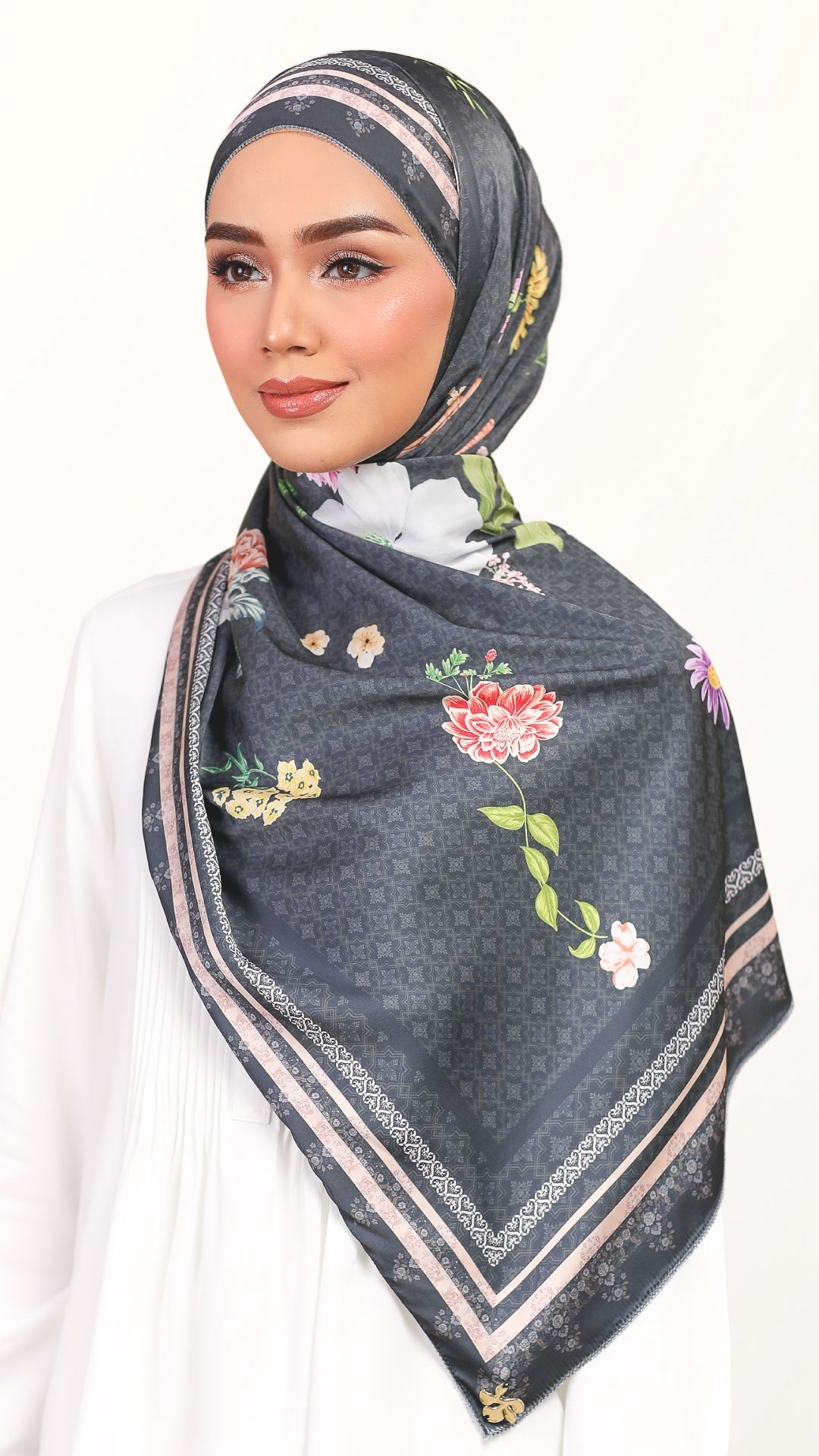 A woman wearing a dark gray patterned hijab with colorful floral prints and beige borders, styled over a white top against a plain white background.