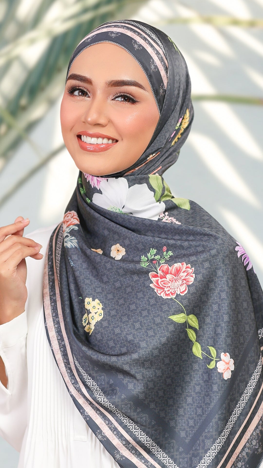 A woman wearing a dark gray patterned hijab with colorful floral prints and striped borders, styled with a white top against a softly blurred outdoor background.