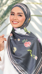 A woman wearing a dark gray patterned hijab with colorful floral prints and striped borders, styled with a white top against a softly blurred outdoor background.