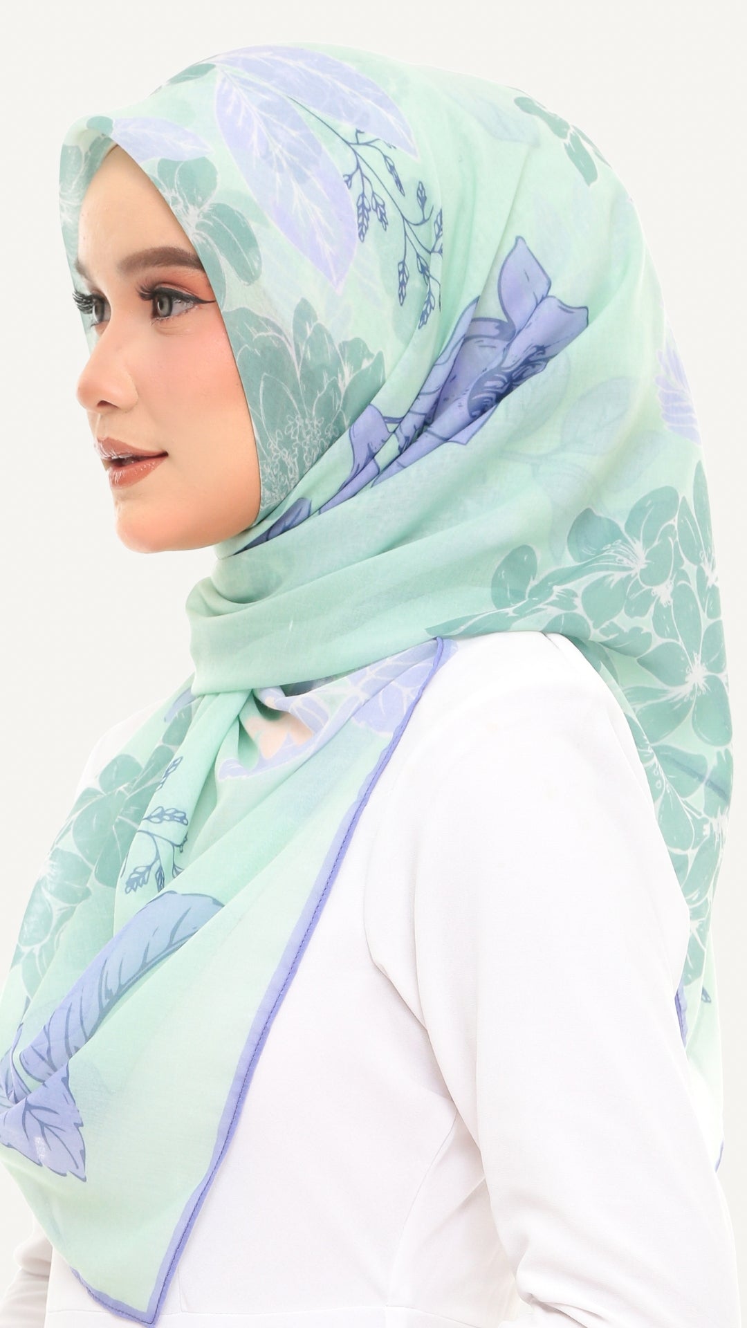 A woman wearing a mint green hijab with a light purple floral pattern, styled over a white top against a plain white background.